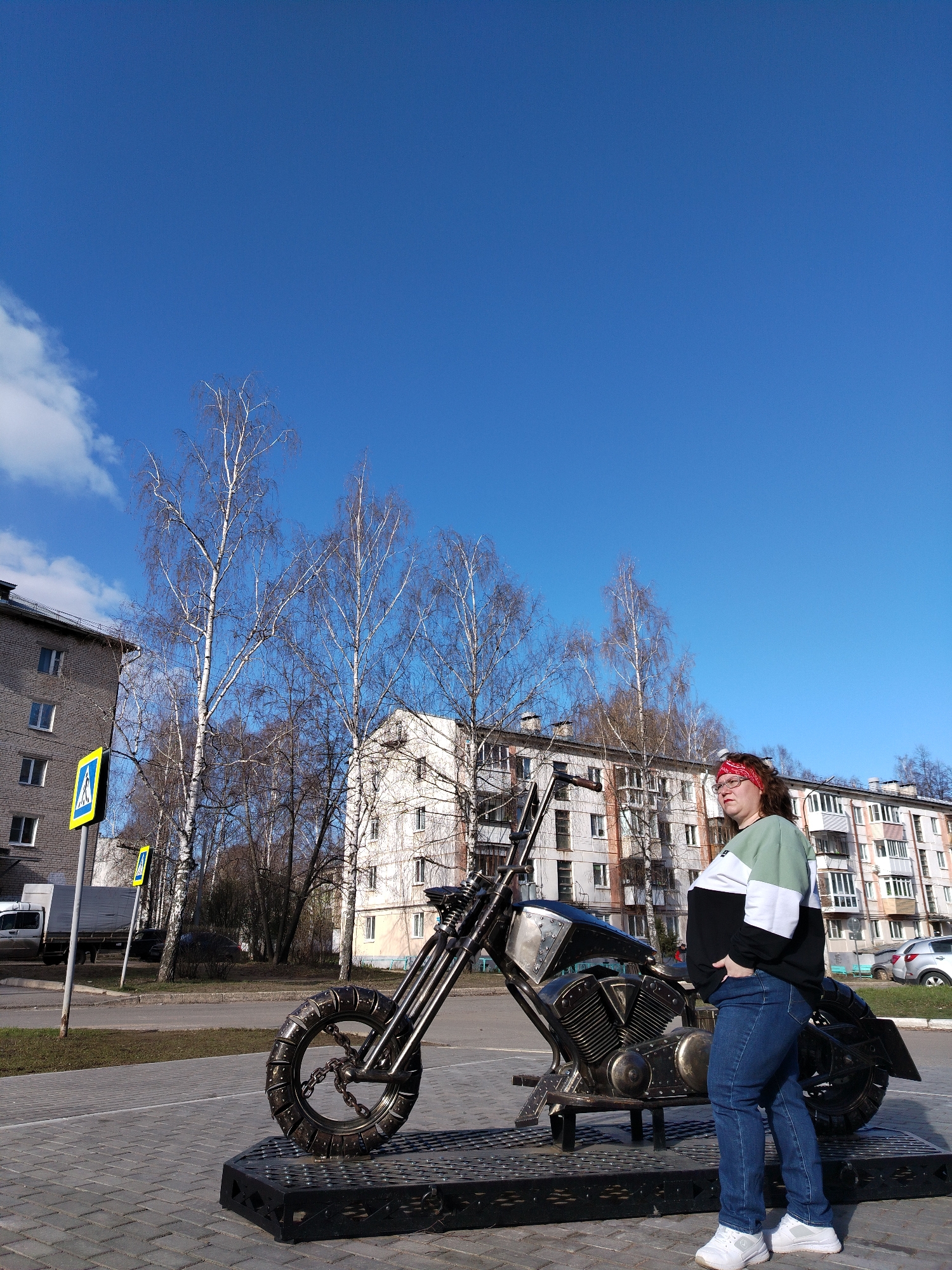 A woman stands on a paved surface next to a metal motorcycle sculpture in Chaikovsky, Russia. She wears a red bandana, a black, white, and green sweatshirt, blue jeans, and white sneakers. The motorcycle is dark-colored and appears to be made of metal. Behind the woman and the sculpture are apartment buildings and bare trees against a blue sky with scattered clouds. A yellow and black pedestrian crossing sign is visible to the left. A white truck and several cars are parked in the background.