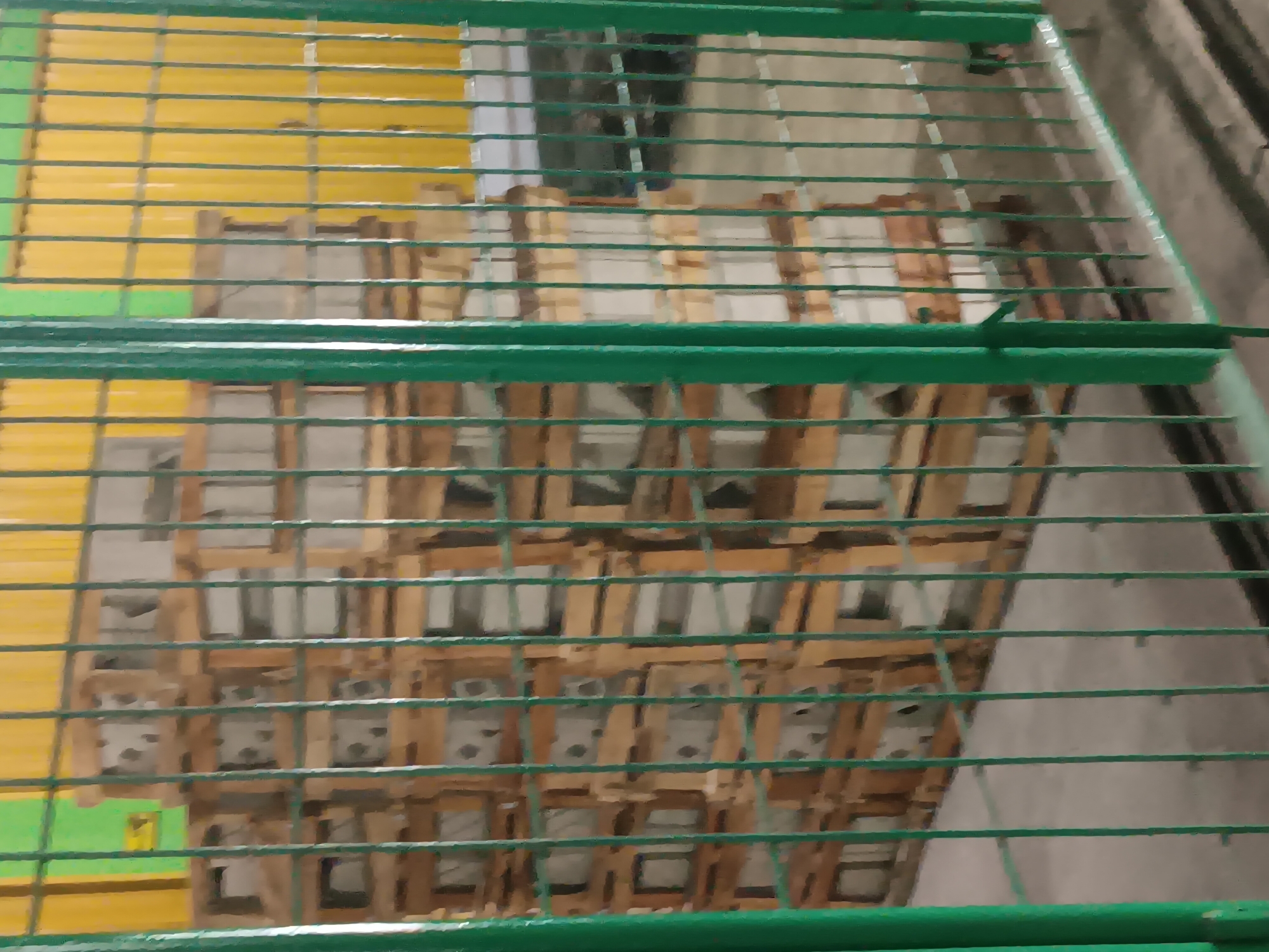 In Batursari, Indonesia, stacks of concrete blocks are stored on wooden pallets behind a green metal fence. The background consists of a corrugated yellow wall and a green vertical panel with a yellow warning sign. The scene appears to be an outdoor storage area or a construction site.