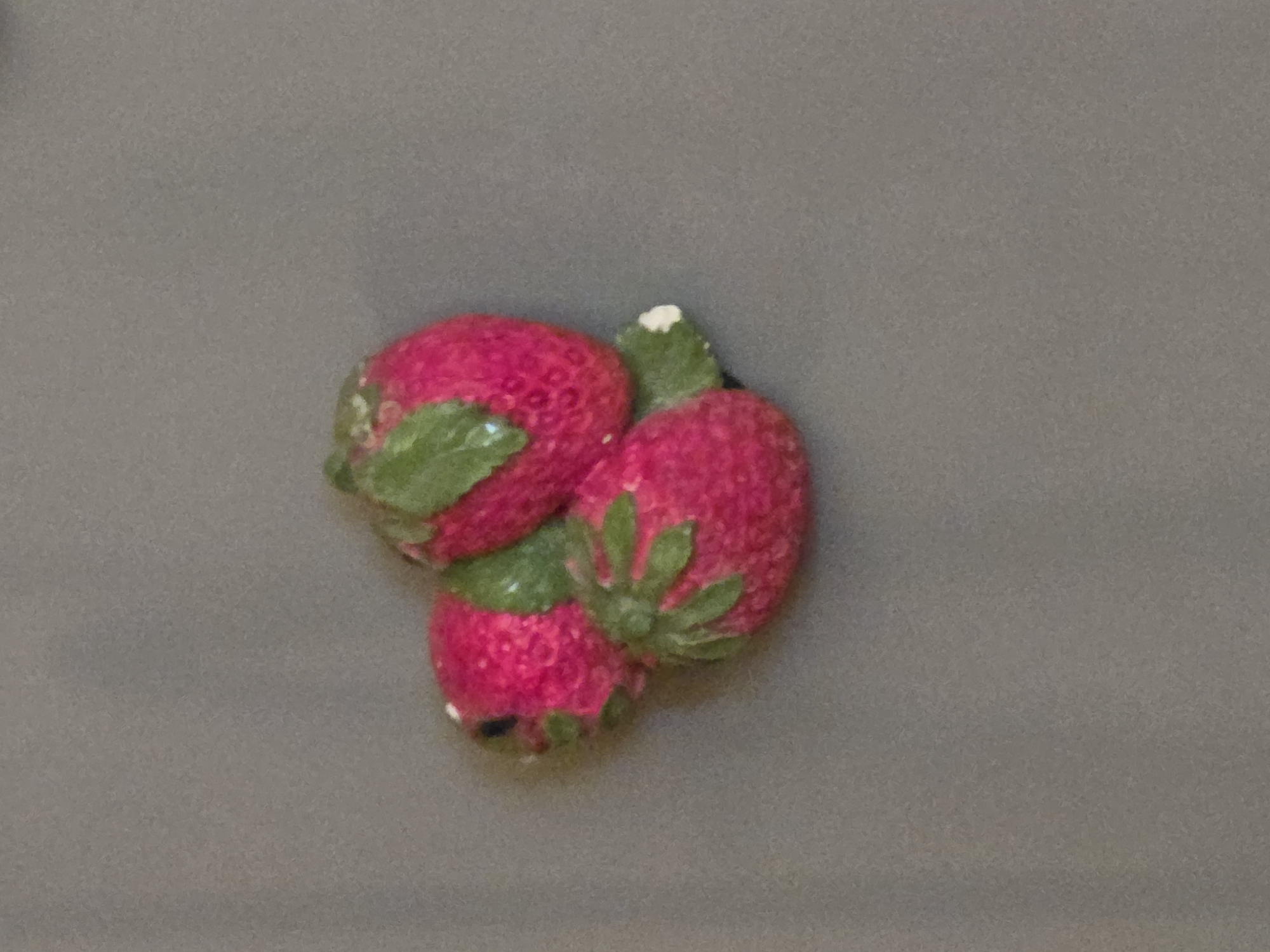 A close-up, high-angle, indoor shot shows a cluster of three pink strawberry-shaped ceramic figures on a gray background. The figures are arranged in a triangular formation, with green leaves adorning each strawberry. The strawberry on the top right is the most prominent, with its stem pointing upwards. The strawberry on the bottom left is partially obscured by the top right strawberry. The third strawberry is positioned between and slightly below the other two. The ceramic figures have a textured surface, with small dots representing seeds. The background is a smooth, matte gray surface. There are no people or any other objects visible in the image. The lighting appears to be soft and diffused, creating subtle shadows around the figures.