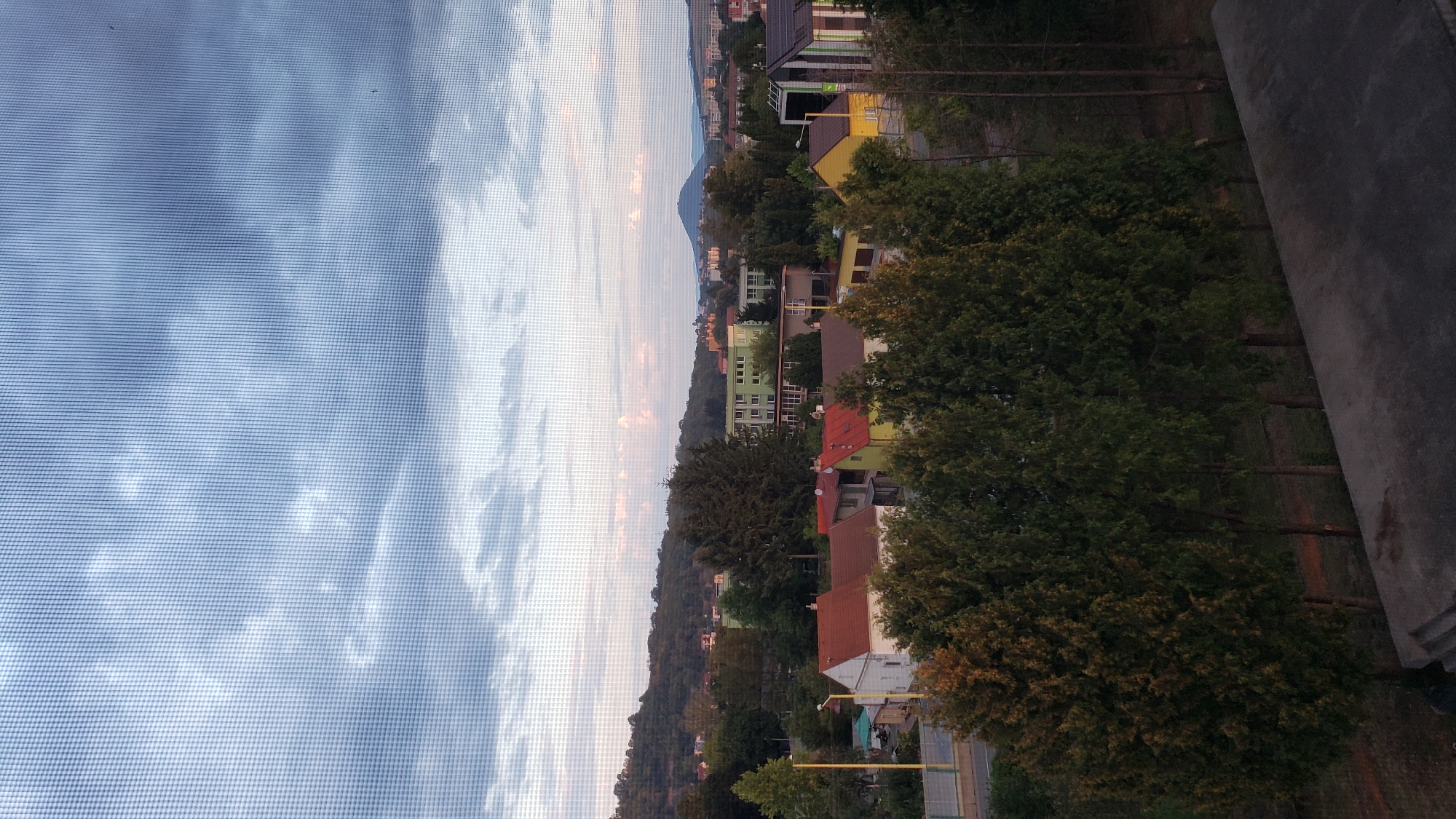 The image depicts a view of a residential area in Prešov, Slovakia, seen through a window screen. The sky is cloudy, with diffused light indicating it's likely late afternoon or early evening. In the foreground, a row of tall, dark green trees partially obscures the view of houses with red and orange tiled roofs. Further back, there are more buildings, including a multi-story green building and other residential structures. In the distance, rolling hills covered in trees form the backdrop, and a distinctive conical mountain is visible on the horizon. The overall impression is a suburban or semi-urban landscape under a muted sky.