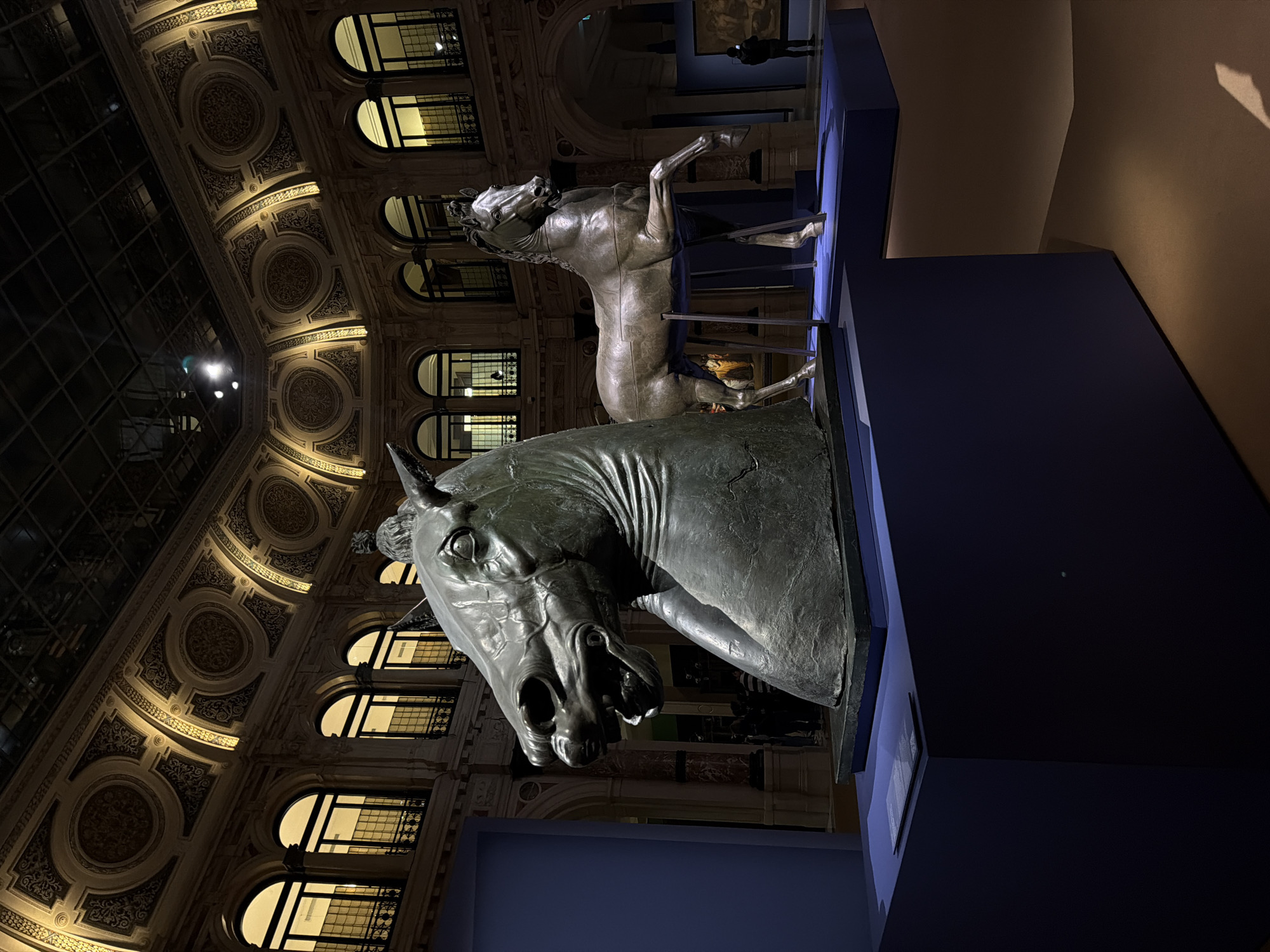 The image depicts an interior view of the Gallerie d'Italia – Piazza Scala in Milan, Italy, featuring an art exhibition. In the foreground, a colossal bronze sculpture of a horse's head, characterized by an open mouth and textured surface, rests on a dark blue, cuboid plinth. A small informational plaque is visible on the plinth to the left of the sculpture. In the mid-ground, a full-body bronze horse sculpture, depicted in a rearing stance with one foreleg raised, stands on a similar dark blue platform, supported by thin vertical metal struts.
The background reveals a grand architectural space with an elaborately decorated, vaulted ceiling featuring gilded or illuminated ornamental patterns and a dark, multi-paned glass skylight. Multiple levels of arched windows with dark frames are visible along the walls, indicating additional interior spaces or upper floors. The lighting is low-key, with strategic illumination highlighting the sculptures and architectural details. Several indistinct human figures are visible in the background; one individual on the far right is clearly observing a framed artwork, while others are partially obscured further back. The floor surface is a dark, uniform brown.