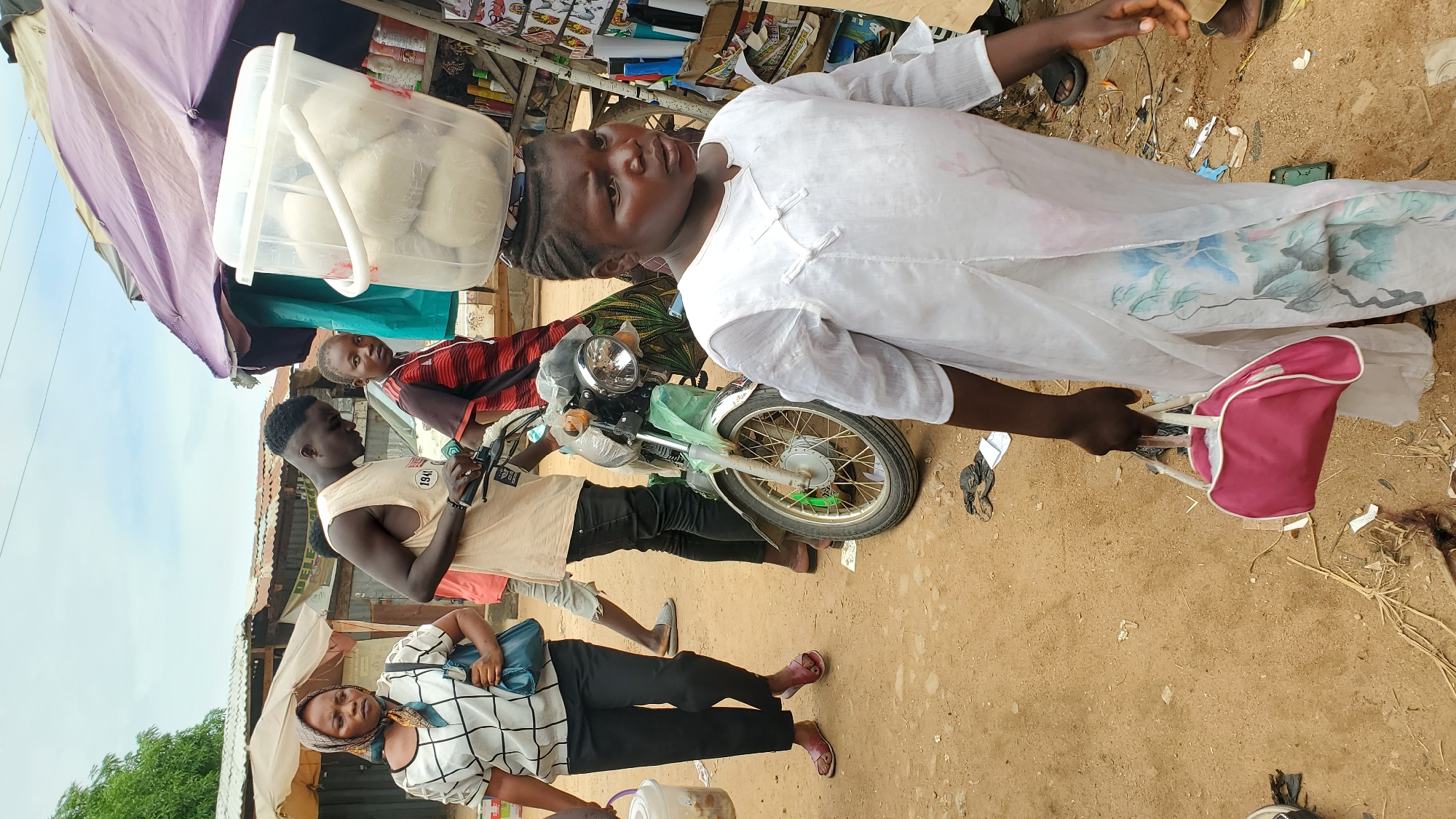 In Jalingo, Nigeria, a woman in a white tunic with floral embroidery and a pink handbag walks forward, carrying a large plastic bucket filled with white food items on her head. To her left, a man in a beige tank top and black trousers is looking down at a green motorcycle, and a woman in a black and white patterned shirt and headscarf is walking away from the camera. Behind the motorcycle, a young person in a red and black striped shirt looks towards the viewer. The scene takes place on a dirt path lined with market stalls under a cloudy sky.