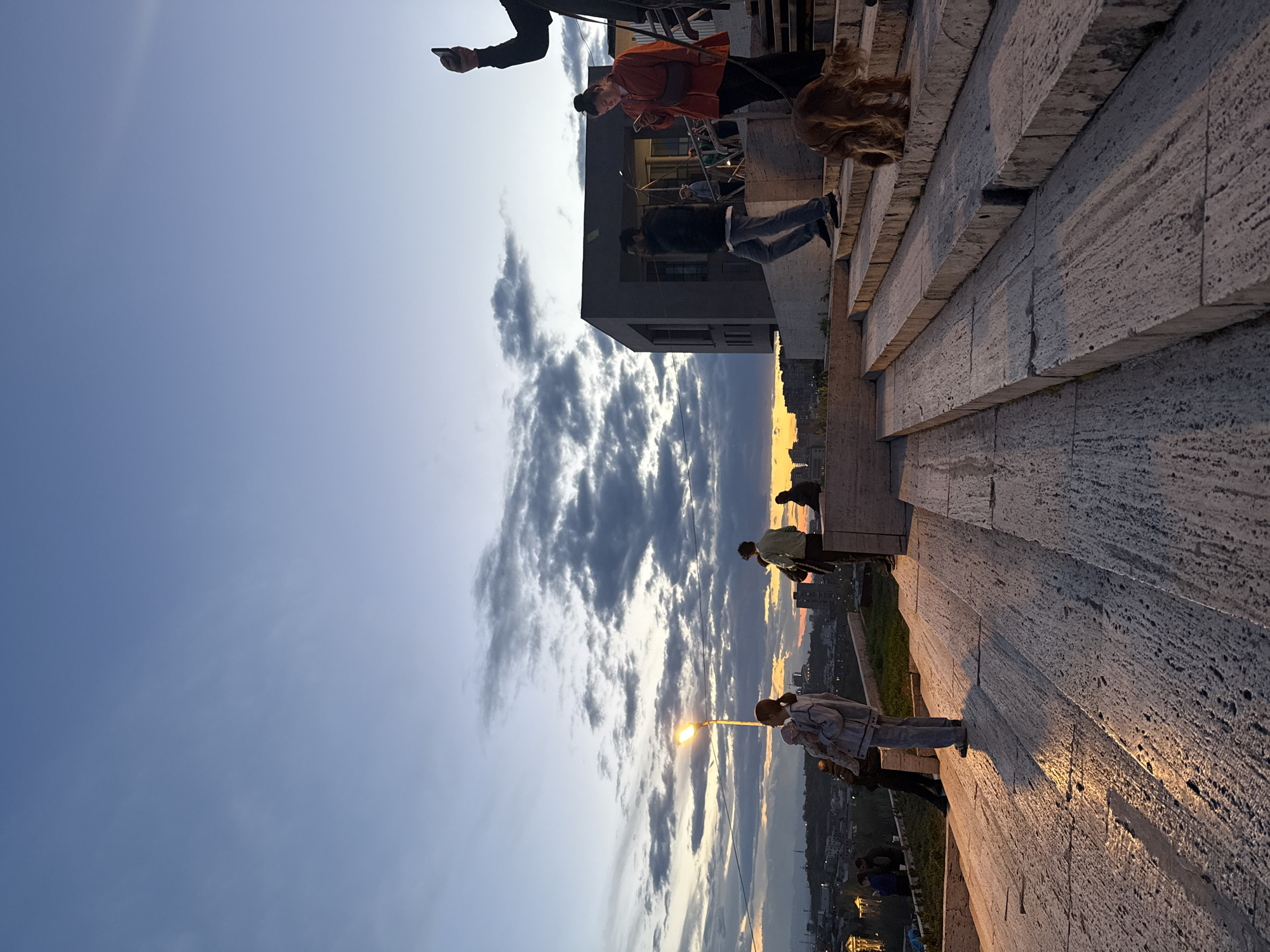 The image, when correctly oriented, captures an outdoor scene at sunset from an elevated architectural structure, likely the Cascade in Yerevan, Armenia. The expansive sky displays a gradient of colors, from a clear light blue overhead to a mix of dramatic, scattered dark grey clouds and vibrant orange and yellow hues at the horizon where the sun is setting. A distant cityscape stretches across the lower part of the horizon.
In the foreground and midground, a series of broad, light-colored stone steps or terraces, textured and possibly made of travertine, descend towards the left. These steps are brilliantly illuminated by the warm, golden light of the setting sun, creating a strong contrast with the cooler tones of the sky and casting long shadows.
Several people are present on the steps and terraces, mostly in silhouette or partially lit. On an upper level, a person wearing an orange jacket and another in a dark jacket and jeans are visible, both appearing to observe the view. A person with distinctive long, reddish-brown hair is seen on one of the steps. Further down, near a brightly lit street lamp, a few individuals stand, one prominent in a light-colored jacket and pants. A hand holding a smartphone is visible in the upper right, suggesting someone is documenting the scenic moment. The activity appears to be casual observation and enjoyment of the sunset over the city. No visible text is present in the image.