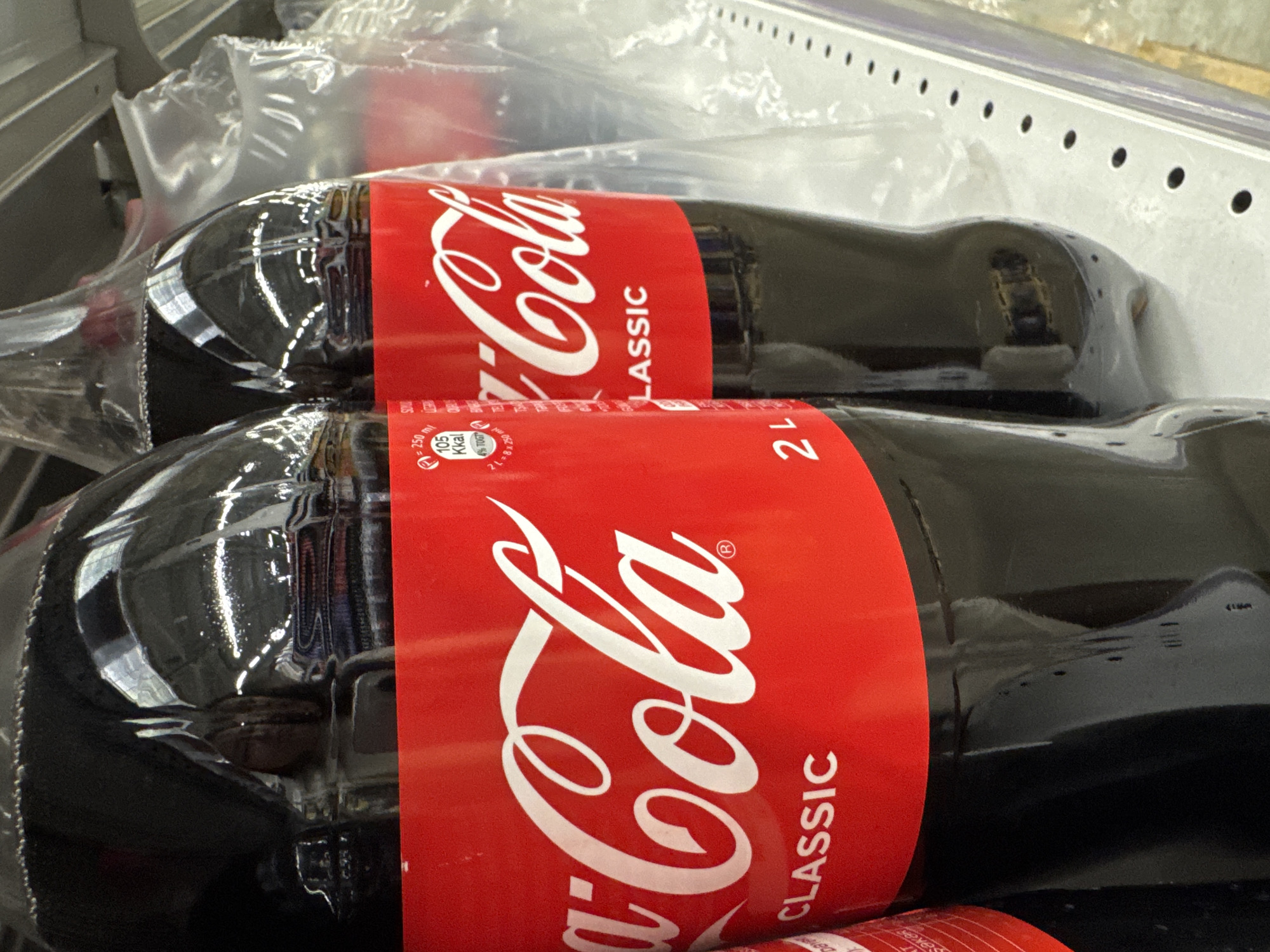 The image shows two bottles of Coca-Cola Classic, packaged together. The bottles are made of dark plastic and are partially filled with a dark liquid. Each bottle has a red label with the white Coca-Cola logo. The label also includes the text "CLASSIC". One of the labels also has nutritional information, showing "105 Kkal" per 250 ml and "0% TOTAL". The bottles are lying horizontally, possibly on a shelf in a store or in a delivery box, given the context of Новомарьевская, Russia. The packaging is clear plastic wrap, also visible. The lighting suggests an indoor environment. No people are visible in the image.