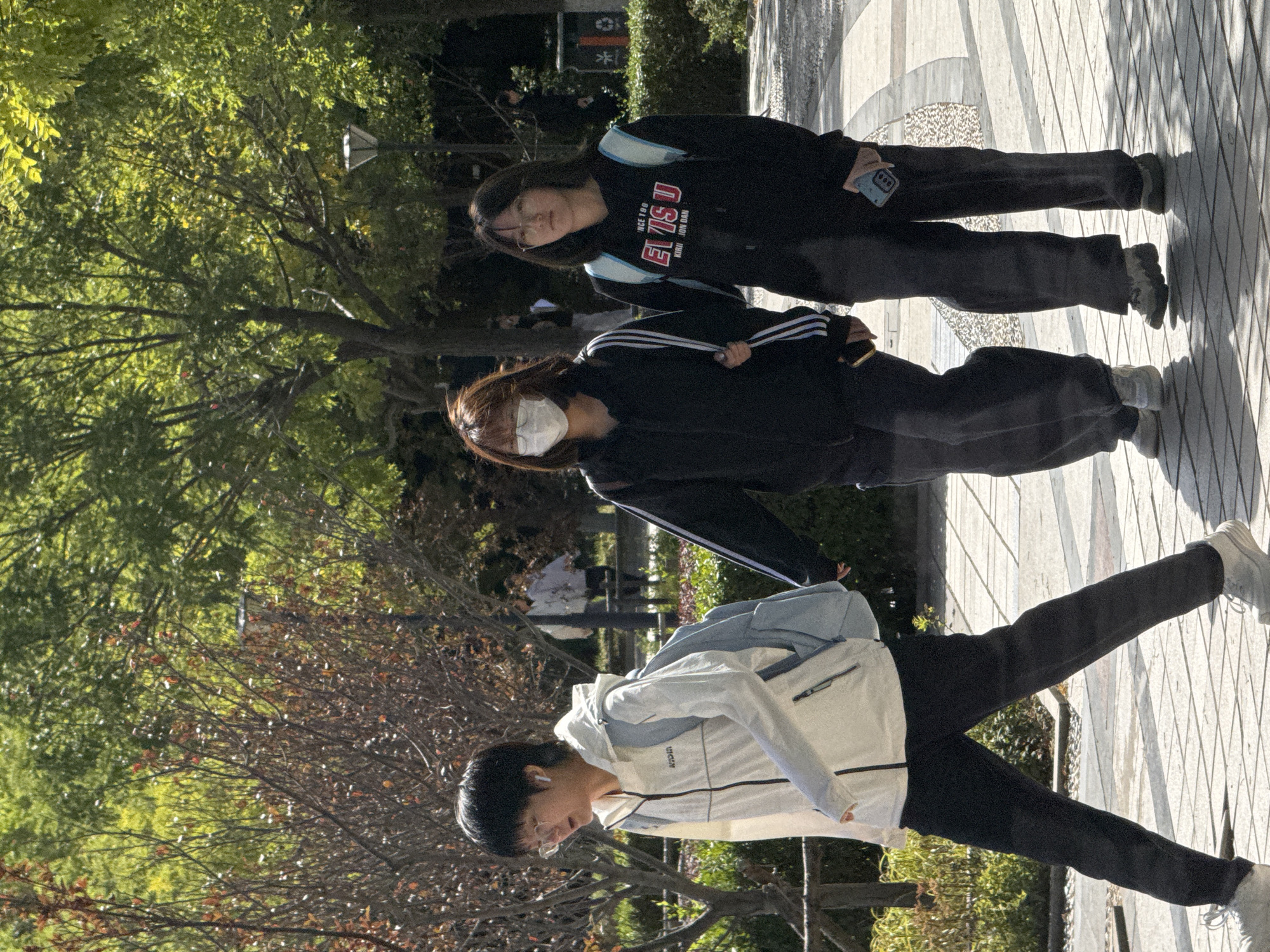 Three young individuals are walking on a paved pathway in a park-like setting, possibly on the campus of a university or a public garden. The foreground features three individuals in motion. The person on the far left, with short dark hair and glasses, wears a white jacket with blue accents and a light blue backpack. They are wearing dark pants and white sneakers. To their right, a person with dark hair, wearing a face mask and a black hooded jacket with white stripes, walks with their arm linked with the person next to them. This individual is also wearing dark pants and light-colored sneakers. The third individual, on the far right, has dark hair and glasses, and wears a black hooded sweatshirt with the text "ELVISU" prominently displayed, along with a light blue backpack. They are wearing dark pants and gray sneakers. Sunlight casts shadows on the pathway, indicating it is daytime. Trees with green foliage and some bare branches surround the pathway. In the background, other individuals are visible walking in the distance, and there is a trash can and lamppost. The overall scene suggests a casual outdoor activity.