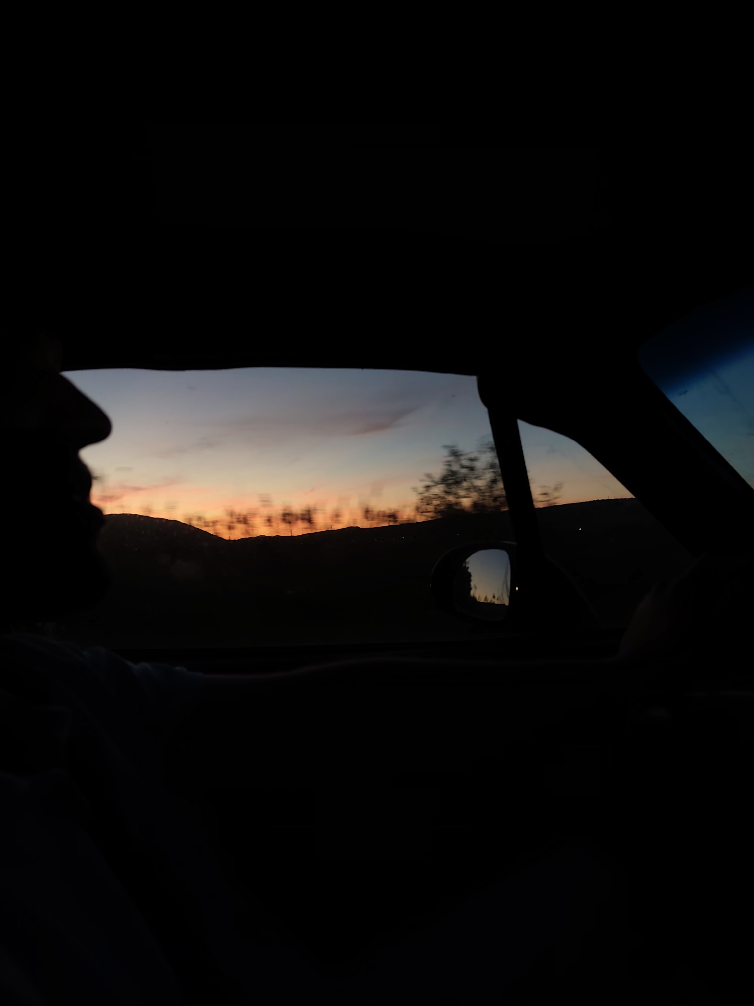 A photograph taken from inside a vehicle at dusk or dawn, showing the silhouette of the driver and the interior of the car frame a landscape through the window. The window view reveals hills and a sky with bands of orange and blue. The side mirror reflects a similar landscape. The location is unknown.