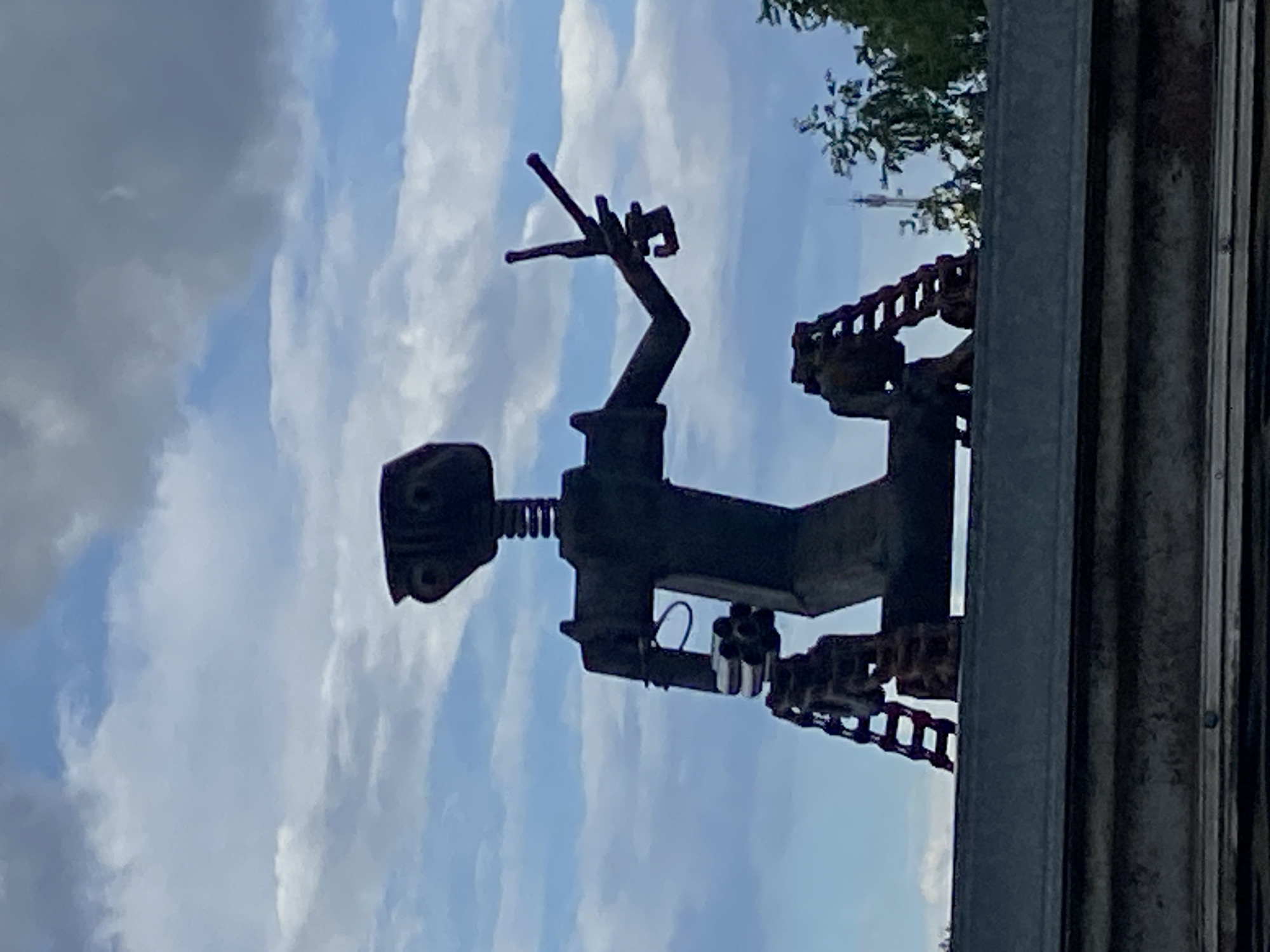 The image shows a metallic sculpture of a robot-like figure. The figure appears to be constructed from scrap metal and industrial parts. It features a head with circular elements, a torso, articulated arms, and chain-like components possibly representing legs or appendages. The sculpture is positioned outdoors, with a cloudy sky visible in the background. Part of a building or structure is visible to the right. A tower in the distance implies that the sculpture is in a city. Given the prompt, this location is Фоки, Russia. The specific location is impossible to ascertain from the given image.
