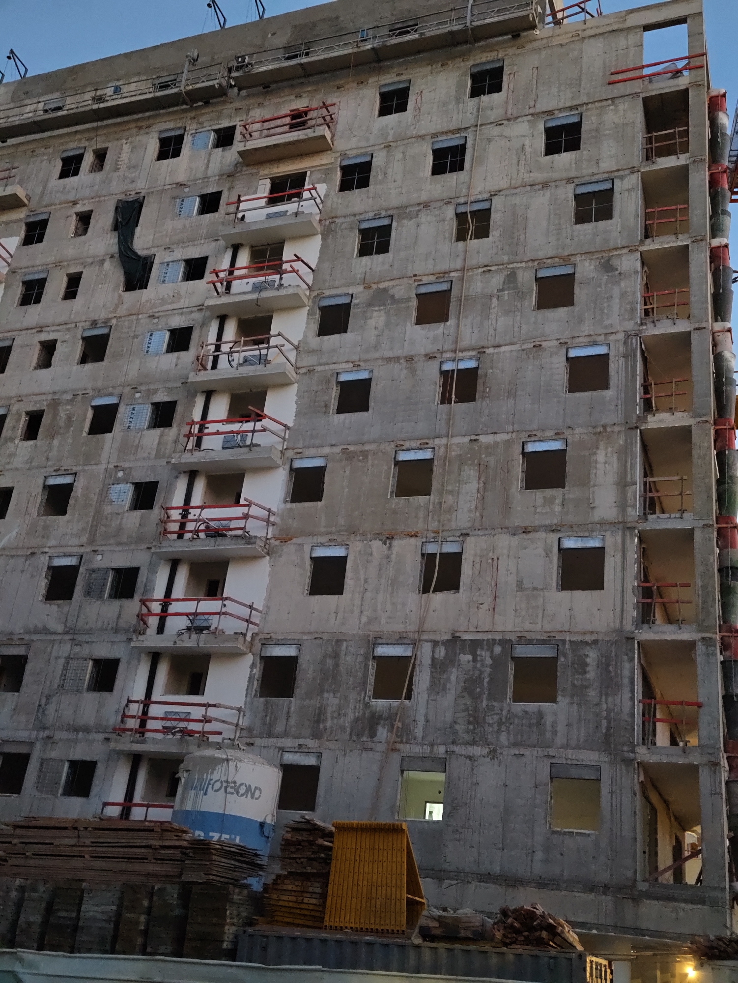 The image shows a multi-story concrete building under construction in Petah Tikva, Israel. The building has a rough, unfinished exterior with numerous openings for windows and balconies. Red railings are visible on some of the balconies, and construction materials, including stacks of wooden planks and a large cylindrical tank with blue and white markings, are present at the base of the building. Scaffolding and construction equipment are also visible. The sky above is a clear, pale blue, indicating daytime. The scene depicts an active construction site.