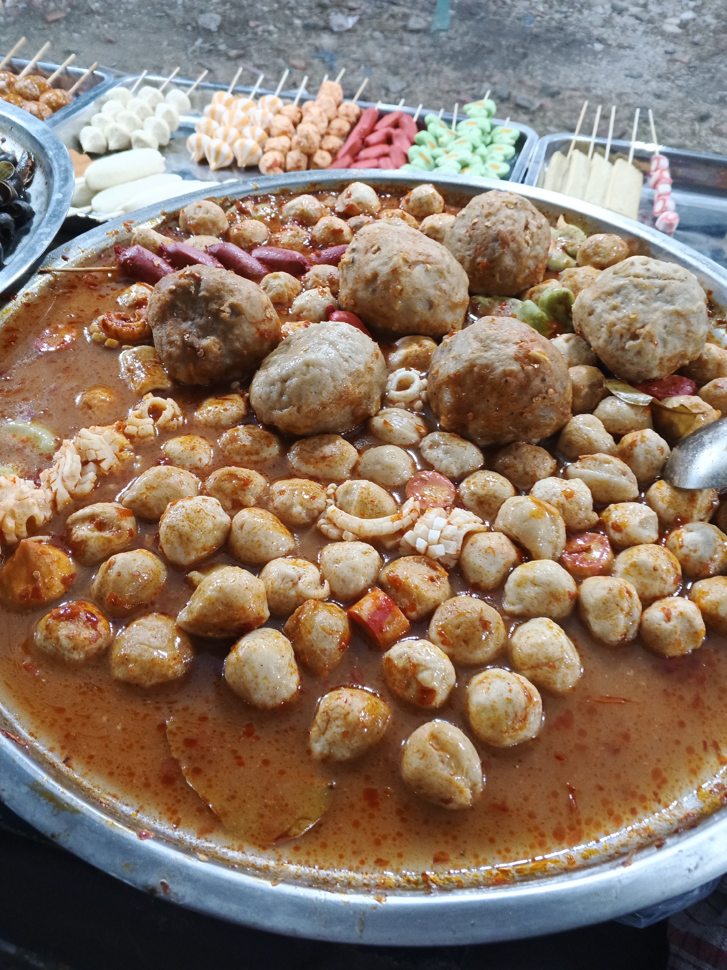 A close-up shot shows a large, round, metal tray filled with various types of meatballs and other food items submerged in a reddish-brown broth. The meatballs vary in size and texture, with some being smooth and round, while others are larger and appear to be more coarsely ground. Several sausages, cut into bite-sized pieces, are also visible amongst the meatballs. Some pieces of what appear to be calamari rings and pieces of red chili peppers are scattered throughout the tray. The broth has an oily sheen and flecks of spices are dispersed within it. In the background, out of focus, are several trays containing rows of skewers with different colored food items, suggesting this is a food stall or market setting. One tray in the upper left corner contains black olives. The overall scene depicts a savory dish being prepared or served, likely from a street food vendor. The image appears to have been taken in a public space, possibly outdoors, given the rough texture of the ground visible in the background. The lighting suggests it could be daytime. There are no people clearly visible in the frame, but the presence of food suggests human activity. The image is a still photograph and captures a moment of food display.