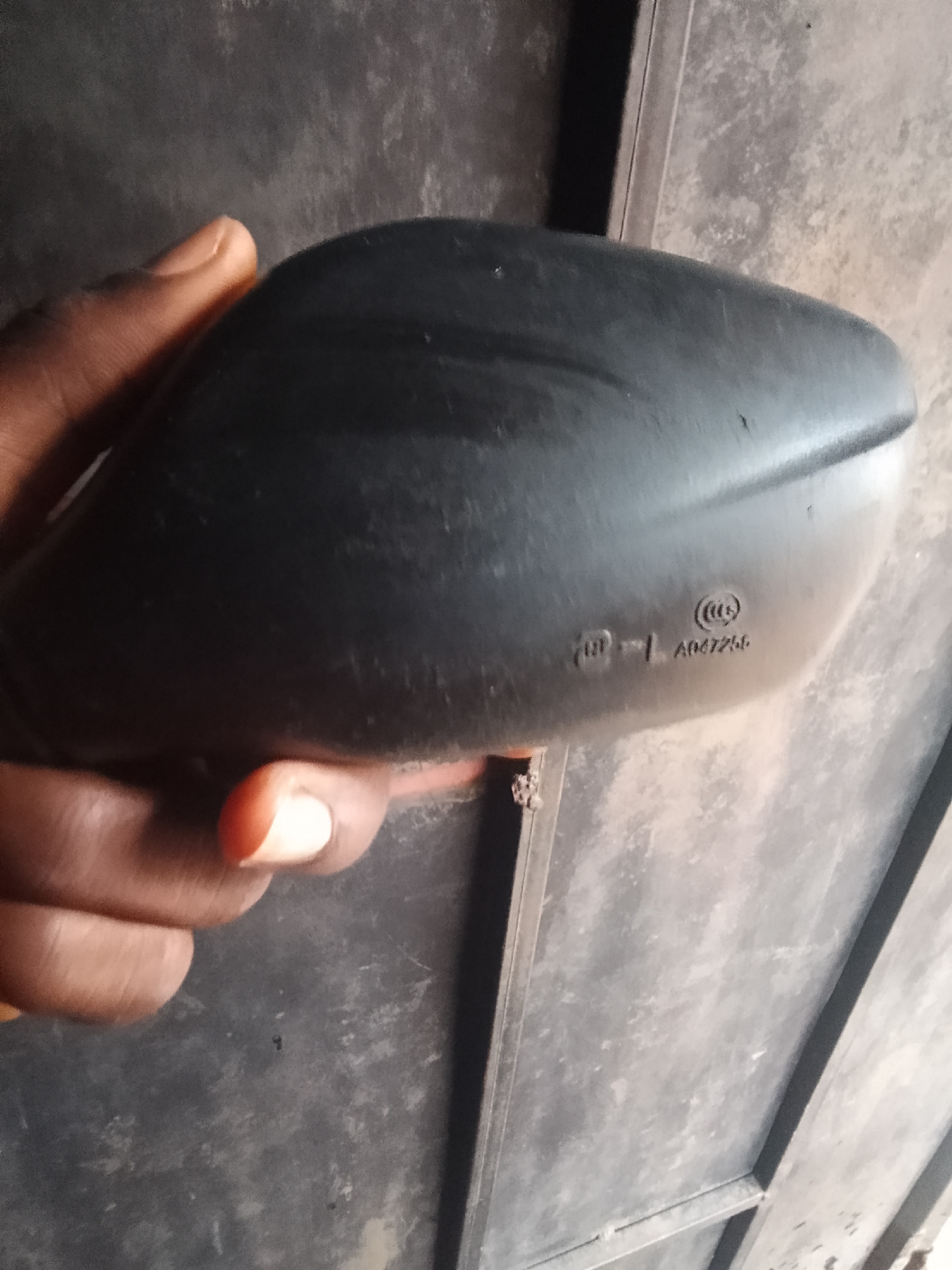 The image shows a close-up view of a black, curved object being held by a hand with dark skin. The object appears to be a car part, possibly a side mirror housing, with some embossed markings on its surface. The markings include a circular symbol and what looks like alphanumeric characters. The background is a textured, mottled surface, possibly a wall or a door, with a muted color palette of grays and browns. The lighting is dim, creating shadows and highlighting the texture of the objects. There is no visible indication of the location being Jalingo, Nigeria, or any specific urban environment.