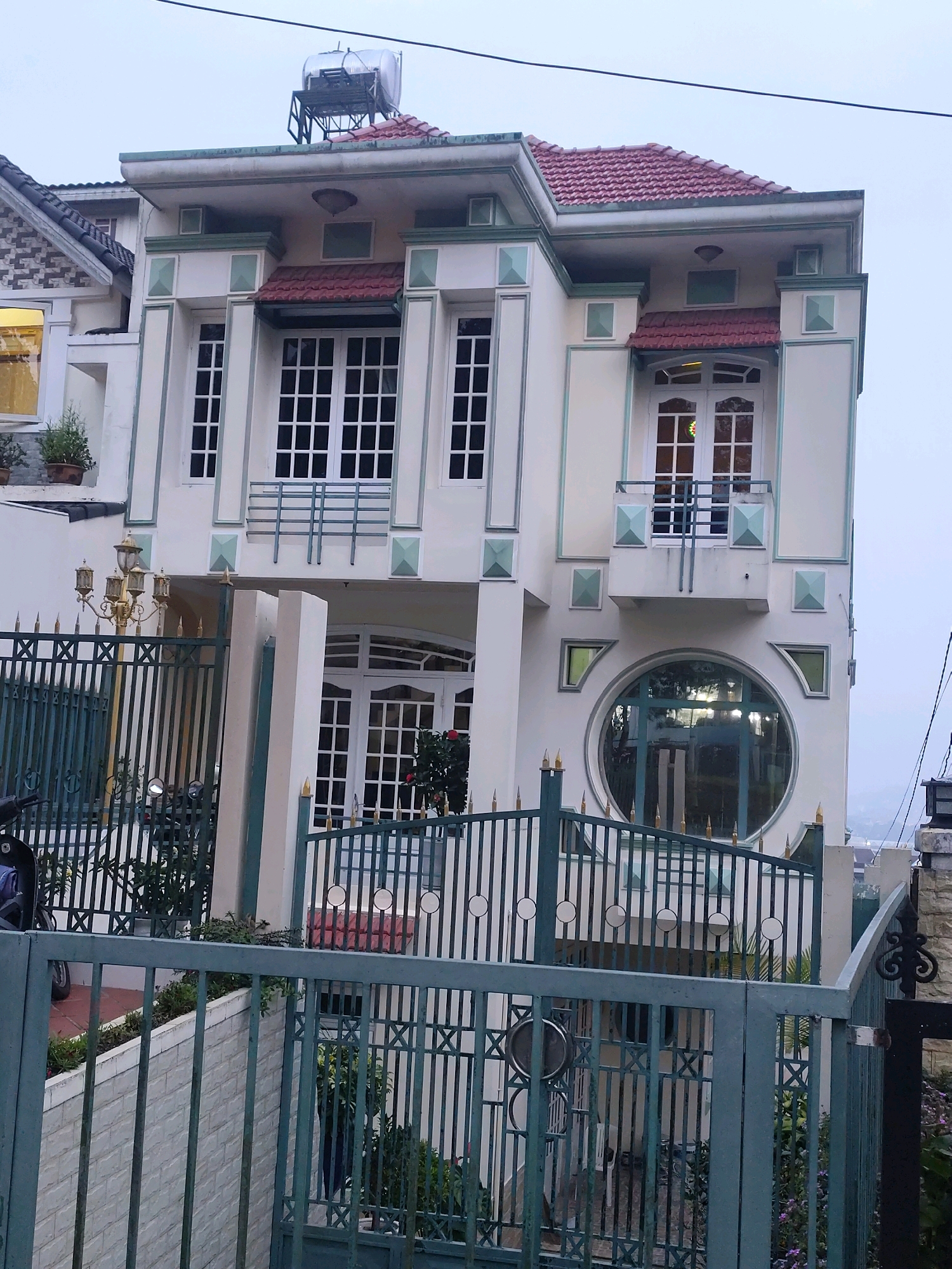 The image shows a multi-story house in Lâm Viên - Đà Lạt, Vietnam. The house has a cream-colored facade with green and white trim. It features several windows, including a prominent circular window on the second floor and multiple rectangular windows with white frames and a grid pattern. The roof is tiled in red. A green metal fence surrounds the property, with a tall, ornate lamppost positioned just inside the gate. Vegetation, including potted plants and bushes, is visible around the house and within the fenced area. The sky is overcast. A motorcycle is partially visible behind the fence. A solar water heater is mounted on the roof.