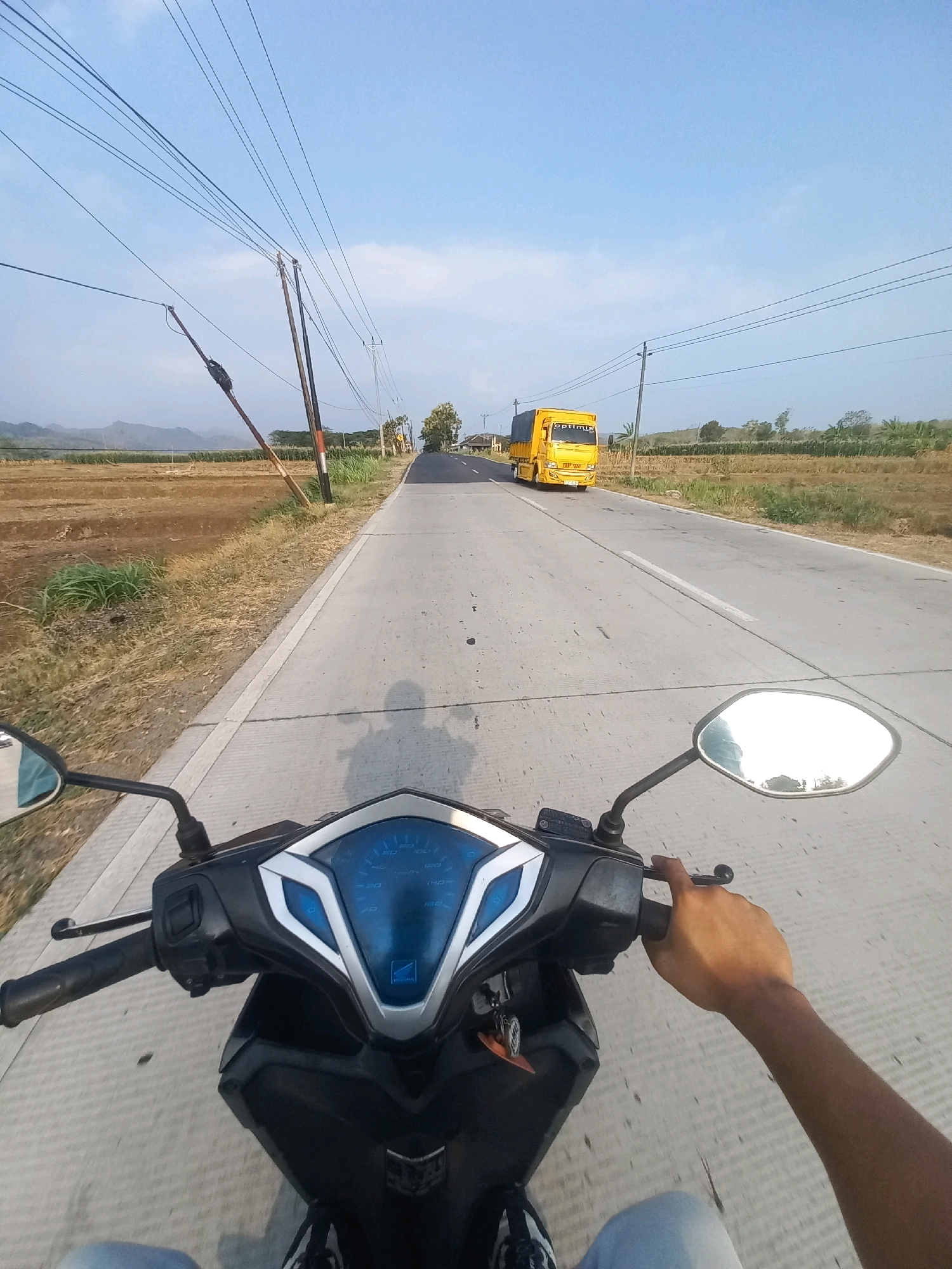 This first-person perspective image captures a motorcycle ride on a paved road in Baturetno, Indonesia. The rider's hands grip the handlebars, and their legs and feet are visible in the lower portion of the frame, suggesting they are wearing light-colored pants and sneakers. The motorcycle's blue-lit dashboard is prominent in the foreground, displaying speed markings up to 160.
The road stretches ahead, with dry, cultivated fields flanking both sides. In the distance, a bright yellow truck with the word "OPTIMUS" visible on its side is driving away from the viewer. Utility poles with numerous wires are scattered along the roadside, indicating the presence of electricity infrastructure. The sky is a clear blue with a few wispy clouds, and a gentle landscape of hills can be seen in the far background. The overall scene depicts a journey through a rural or semi-rural Indonesian landscape.