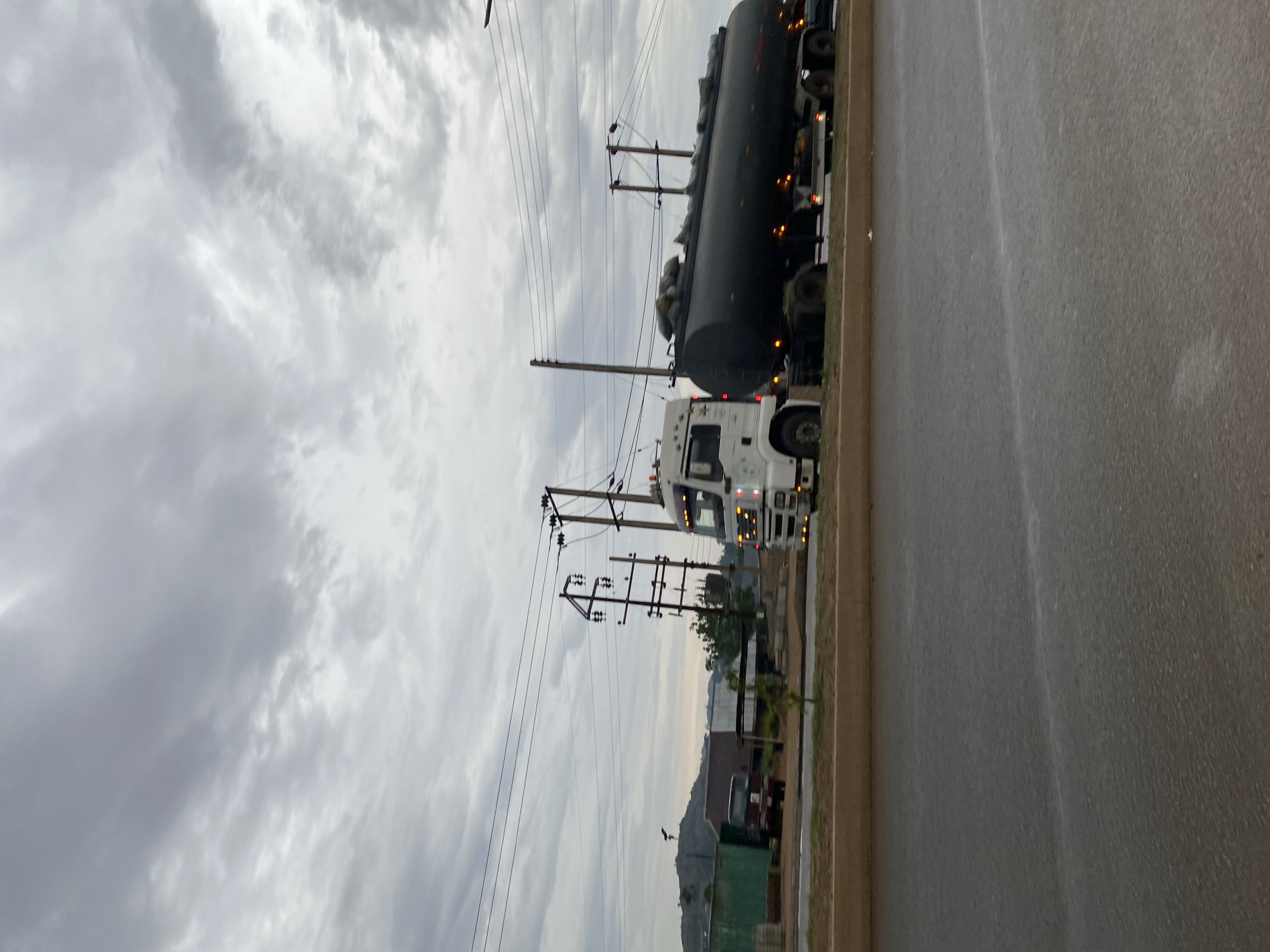 The image, oriented horizontally, depicts a roadside scene under an overcast sky. A prominent white truck acts as a tractor unit, pulling a large, black, cylindrical tank trailer. The trailer appears to be carrying a large container or multiple items, possibly for bulk transport of liquids or gases, with some bundled materials or tarps visible on its top. The truck's headlights and amber marker lights are illuminated.
Along the left side of the road, numerous utility poles stand, crisscrossed with a dense network of power lines, receding into the distance. In the midground, behind the utility poles, distant hills or mountains are visible under a hazy sky. To the right of the main truck, parts of several other trucks or vehicles are discernible along the edge of the paved road. The road itself appears dark and possibly damp, with a visible curb separating it from the shoulder where the trucks are situated.
The sky is heavily clouded and overcast, suggesting a dull daytime light without direct sunlight. There are no people visible, and no discernible activity other than the implied transport function of the trucks. No legible text is present on any of the vehicles or structures. The scene conveys a functional, industrial-like setting, likely in a rural or semi-urban area.