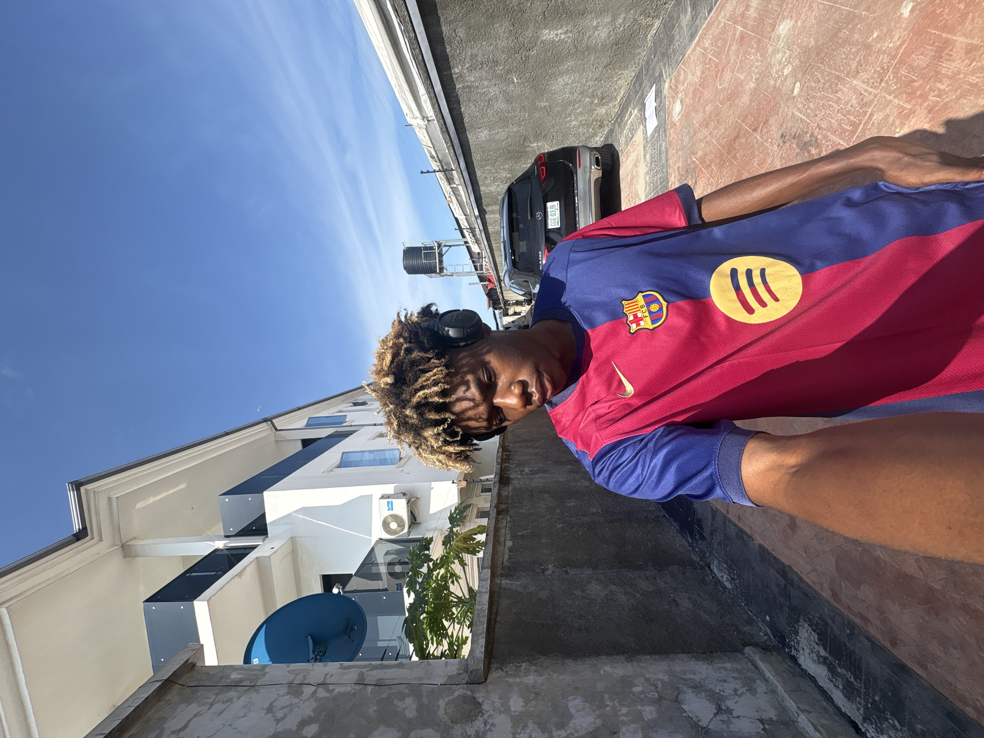 The image depicts a young dark-skinned male taking a selfie outdoors in Igboefon, Nigeria, under a bright, clear blue sky with faint wispy clouds. The subject, positioned in the lower center, wears black over-ear headphones and a red and blue striped FC Barcelona soccer jersey featuring a gold Nike swoosh on the right chest, the FC Barcelona crest on the left chest, and a yellow Spotify logo below it. His hair is styled in light brown and blonde dreadlocks or twists, and he has a nose piercing on his left nostril. He is looking downwards and slightly to his left, with his right arm extended towards the camera, indicating he is holding the device.
To the left and behind the subject, a light-colored multi-story building features dark-colored balcony railings, a large blue satellite dish mounted on its exterior, and an outdoor air conditioning unit. Green foliage, possibly from a papaya tree, is visible between a concrete retaining wall and the building. This textured concrete wall runs horizontally through the mid-ground, separating the foreground from the background structures.
To the right of the subject, a paved area leads to a parked dark-colored SUV, identifiable by its Nigerian license plate "GGE 03JB." Behind the SUV, a metal framework supports a black water tank. The ground in the foreground is a reddish-brown paved surface, possibly interlocking concrete pavers or stained concrete, with a piece of white paper resting on it to the right. The scene is brightly illuminated by direct sunlight, casting distinct shadows.