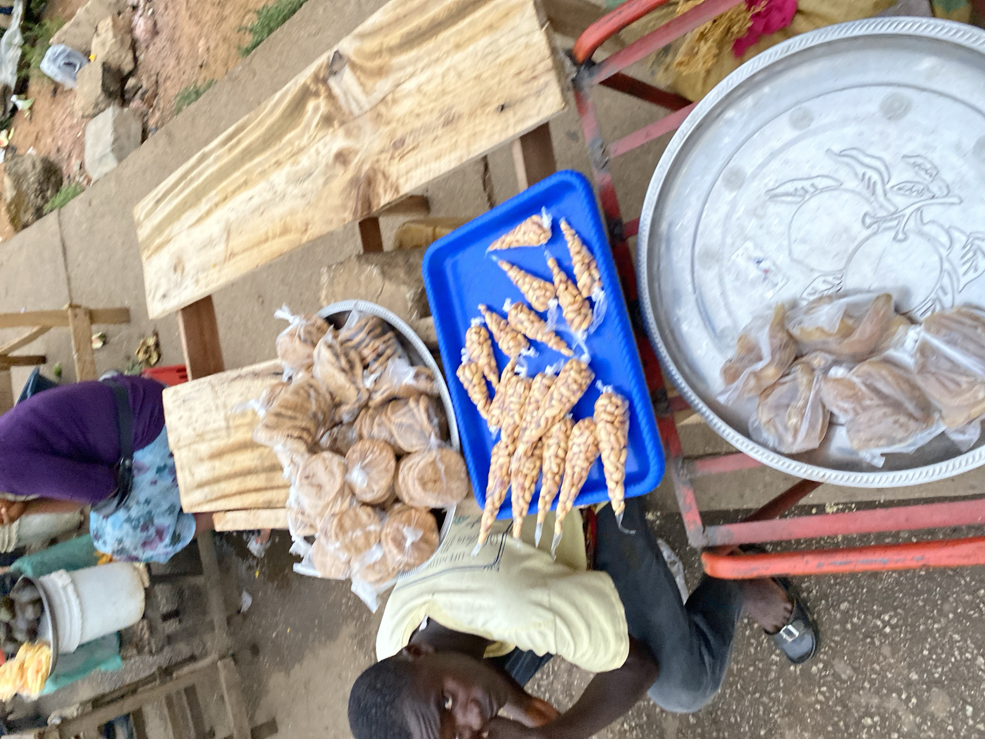 A high-angle shot captures a street vendor in Bukuru, Nigeria, displaying an array of packaged snacks for sale. The vendor, a young Black man wearing a light yellow shirt and dark trousers, is situated amidst his merchandise. His face is partially obscured by his arm, suggesting a moment of pause. A bright blue plastic tray holds cone-shaped packages, likely containing peanuts. To the left, a silver tray contains disc-shaped treats individually wrapped in plastic, while another silver tray displays larger, similarly wrapped items. The goods are arranged on a wooden table and a red metal cart, indicative of a makeshift market stall. The setting is a street scene with uneven pavement and scattered debris, suggesting a busy, urban environment.