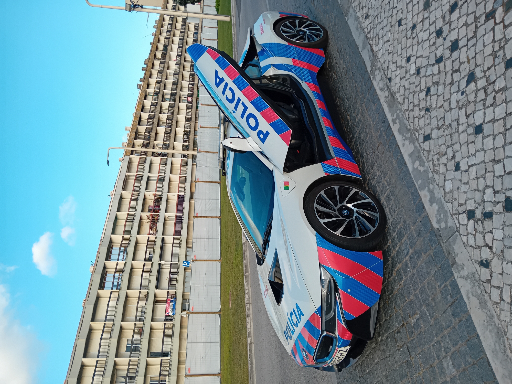 A white BMW i8 police car with the word "POLÍCIA" in blue lettering on its doors is parked on a cobblestone street in Figueira da Foz, Portugal. The car has red and blue stripes on its body. Its butterfly doors are open. The backdrop features a tall, rectangular apartment building with multiple balconies. The sky is blue with a few white clouds. Street lamps are visible.