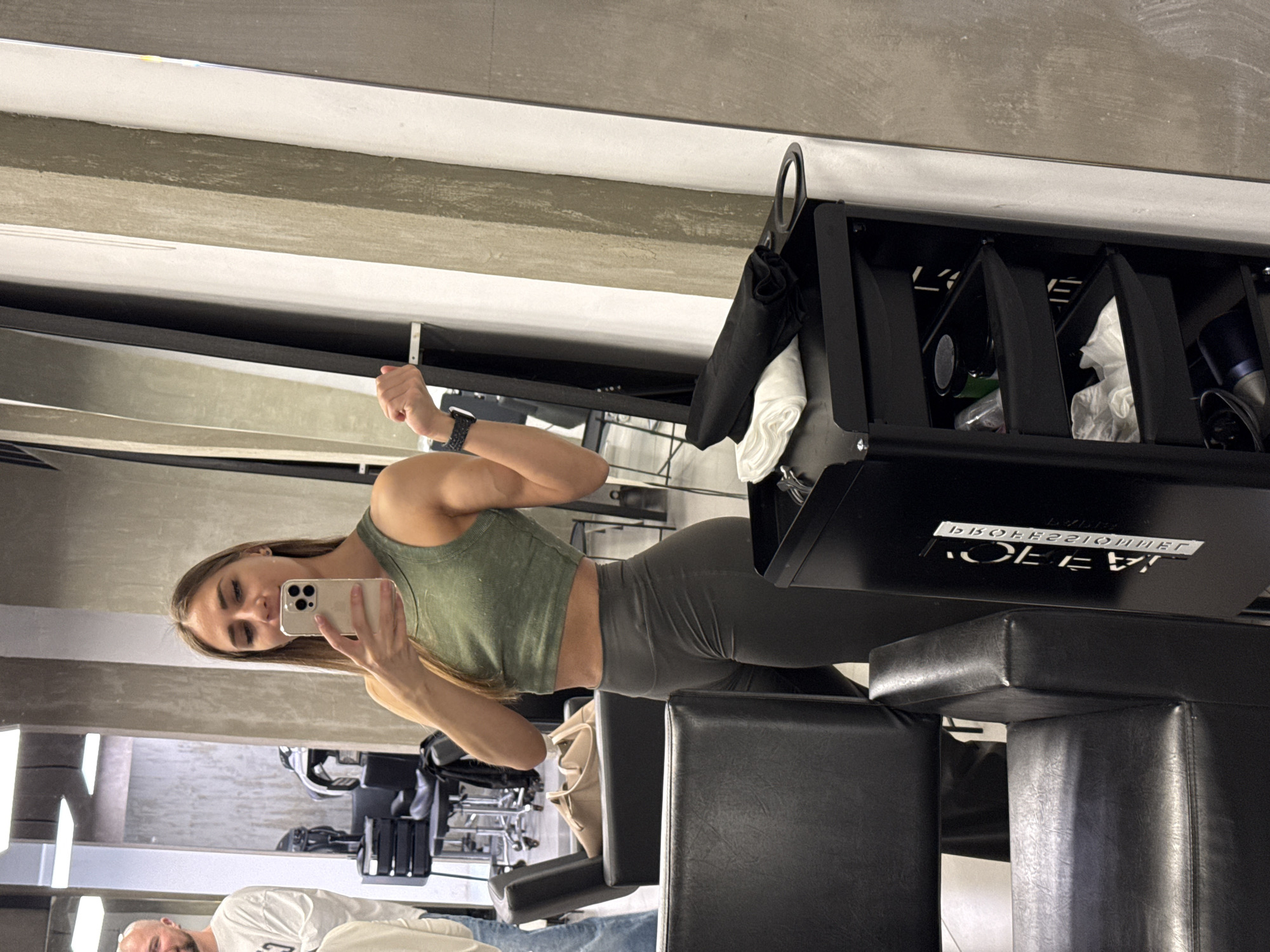 A young woman in an olive-green tank top and grey leggings is taking a selfie in a mirror. She's holding a silver iPhone. Her right arm is flexed. Behind her are black leather gym benches and a black metal storage unit. Below her reflection is a man in a white shirt, only partially visible. The gym setting includes exercise equipment in the background. The upside-down text on the storage unit reads "PROFESSIONAL." There is no information to determine the location of the image in Saint Petersburg, Russia.