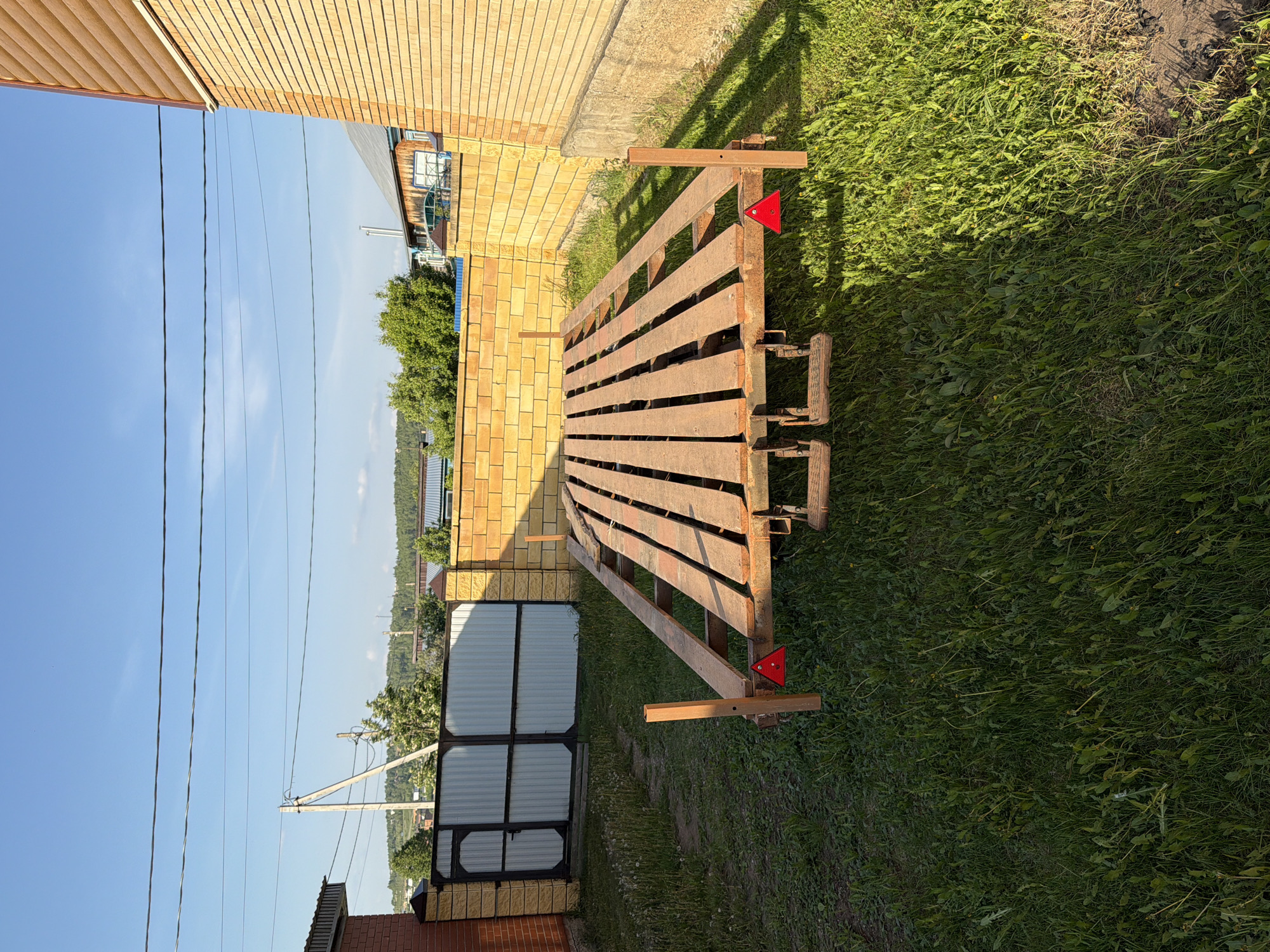 The image depicts a rural scene in Bolsheustyikinskoye, Russia. A small, rusty, open-platform trailer is parked on a patch of green grass. The trailer is oriented with its longer side parallel to a beige brick wall. A smaller fence gate is visible next to the wall. Overhead power lines stretch across a blue sky.