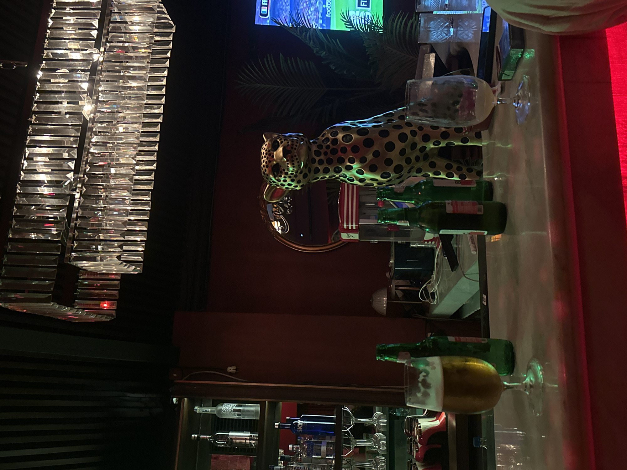 The image depicts an interior scene, likely a bar or nightclub. A crystal chandelier hangs from the dark ceiling. In the mid-ground, a golden leopard statue with black spots is positioned behind a bar. The bar top is a polished, light-colored surface with several objects on it, including glasses, beer bottles, and a pack of cigarettes. In the foreground, a glass of beer sits on the bar. To the right, a large television screen displays a sports match. The walls are a deep red. Shelves filled with various bottles, likely alcoholic beverages, are visible to the left. The overall ambiance appears to be dimly lit with some accent lighting. There is no visible indication of the location being specifically North Kuta, Indonesia.