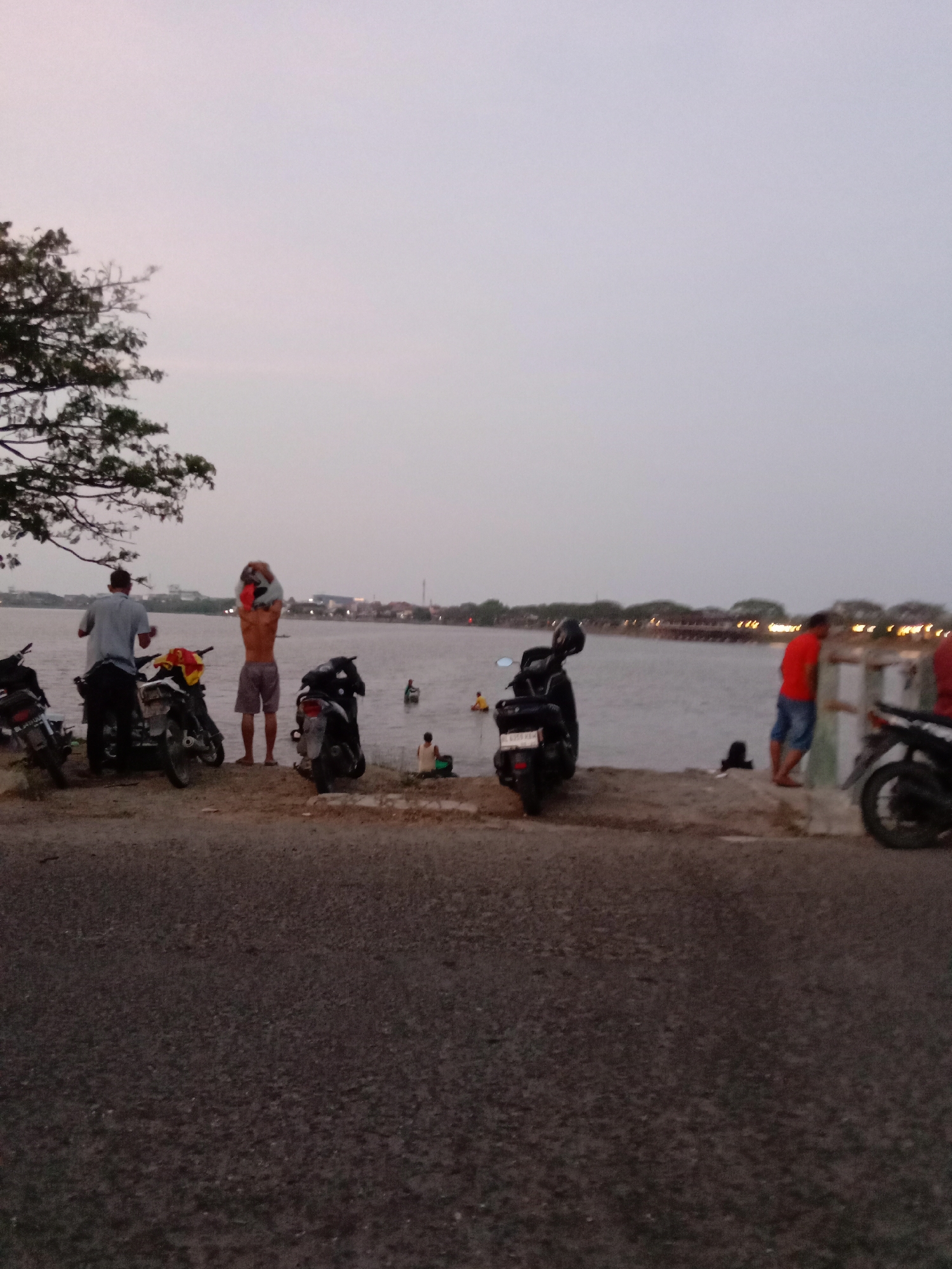The image depicts a waterfront scene in Lhokseumawe, Indonesia, likely near a lake or bay. Several people are present: one man near two motorcycles is raising a helmet over his head, another stands by another motorcycle, and others are at the water's edge, some standing and others sitting. A person is in the water. Several motorcycles are parked near the water's edge on a paved area or road. In the background, a structure possibly a bridge or building with lights is visible across the water. The sky is overcast. A tree is on the left side of the frame.