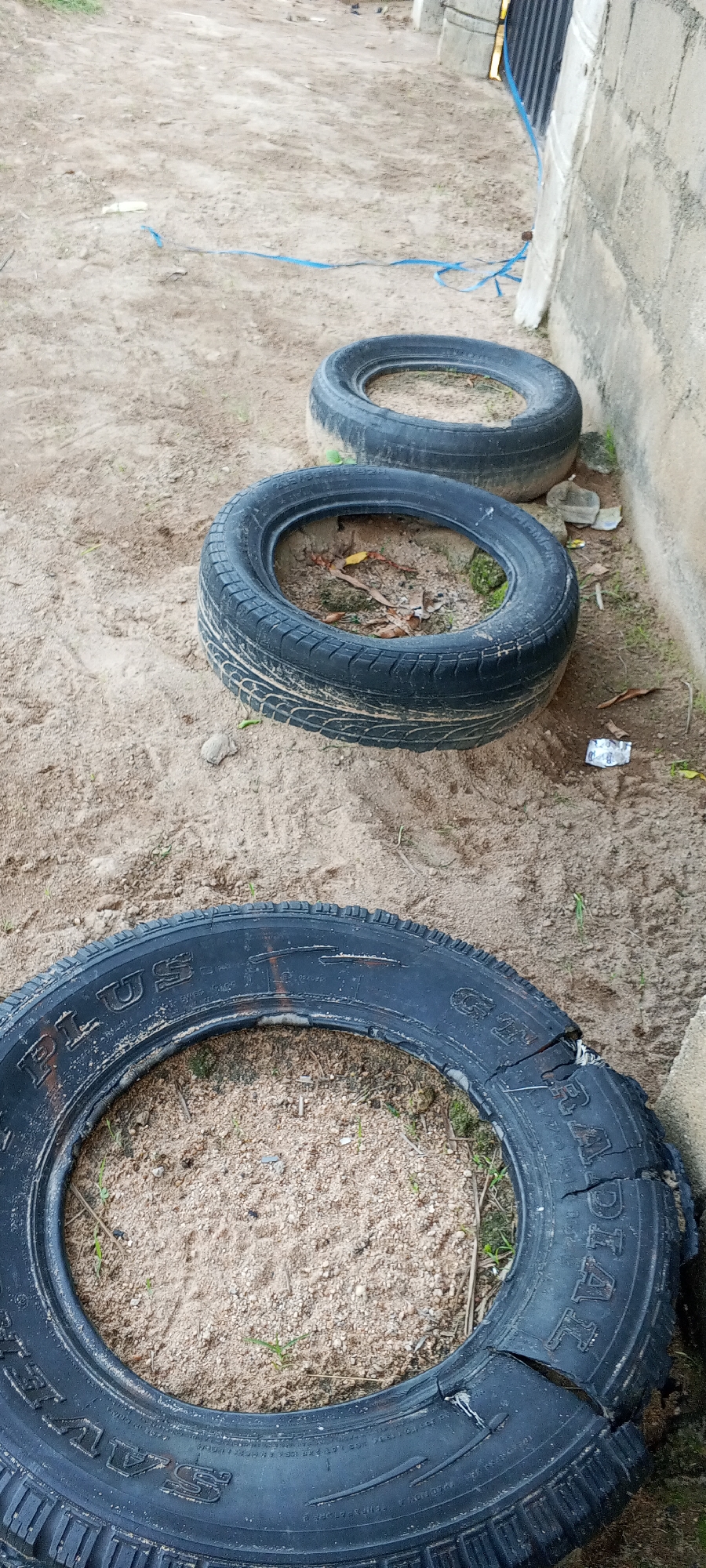 Three used tires are arranged in a line on sandy ground near a cinder block wall. The tires are black and appear to be old, with some signs of wear and tear, particularly on the outermost tire which has a noticeable tear. The ground is sandy with sparse patches of dry grass and some scattered debris, including a torn piece of silver foil packaging. A thin blue cord runs diagonally across the scene in the background. The setting appears to be an outdoor area, possibly a backyard or a less maintained space within a residential or commercial area in Jalingo, Nigeria. The lighting suggests it is daytime. There are no people or animals visible in the image.