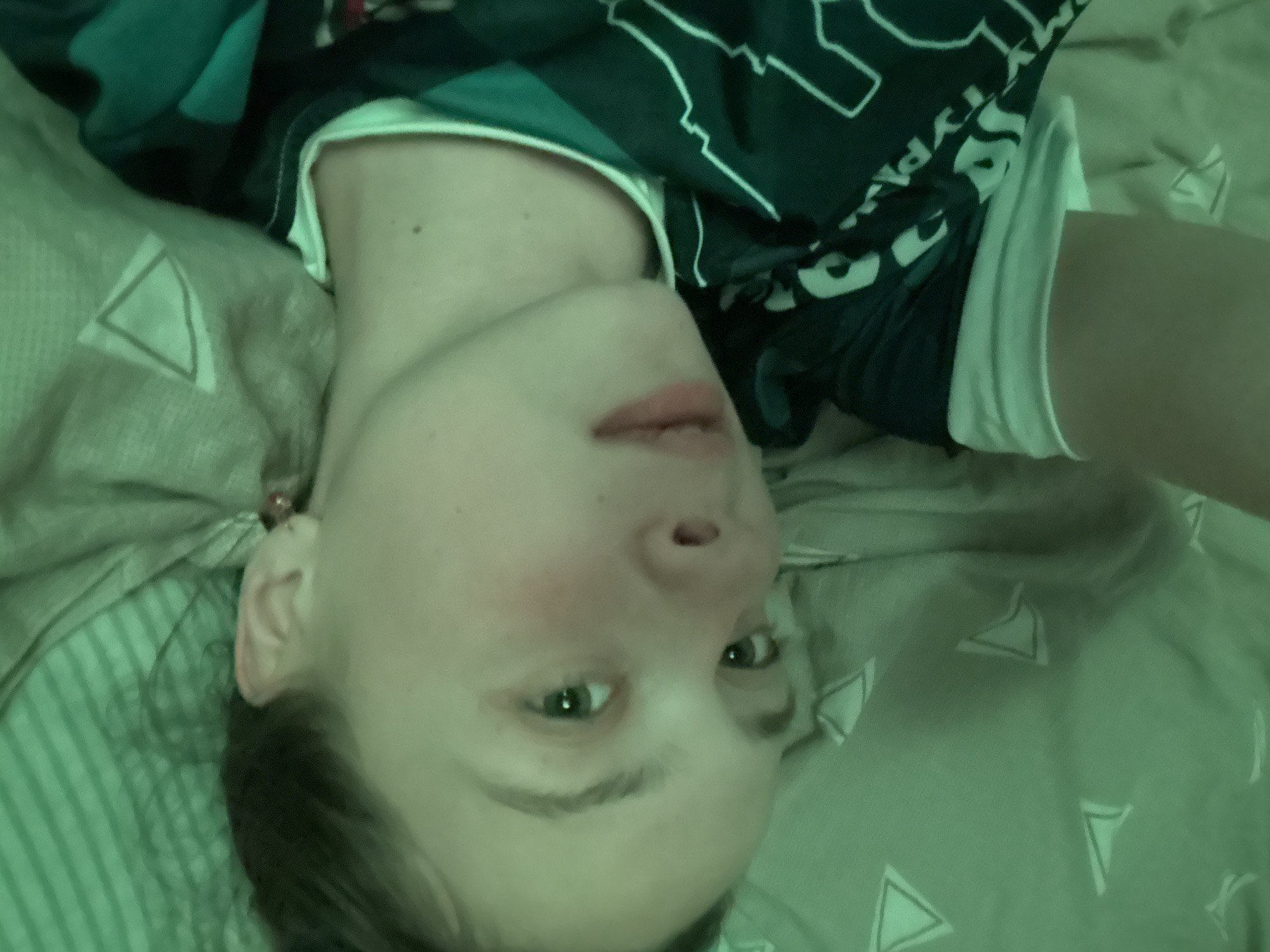 A person lies on a bed in what appears to be a room. The person is wearing a shirt with green and white geometric shapes, and their head is turned upward in the image, which is upside down. The bed linens have a light green and gray color with triangular designs. The lighting in the room seems dim and with a greenish tint. The location is Прикамский, Russia, although this cannot be confirmed from the image.