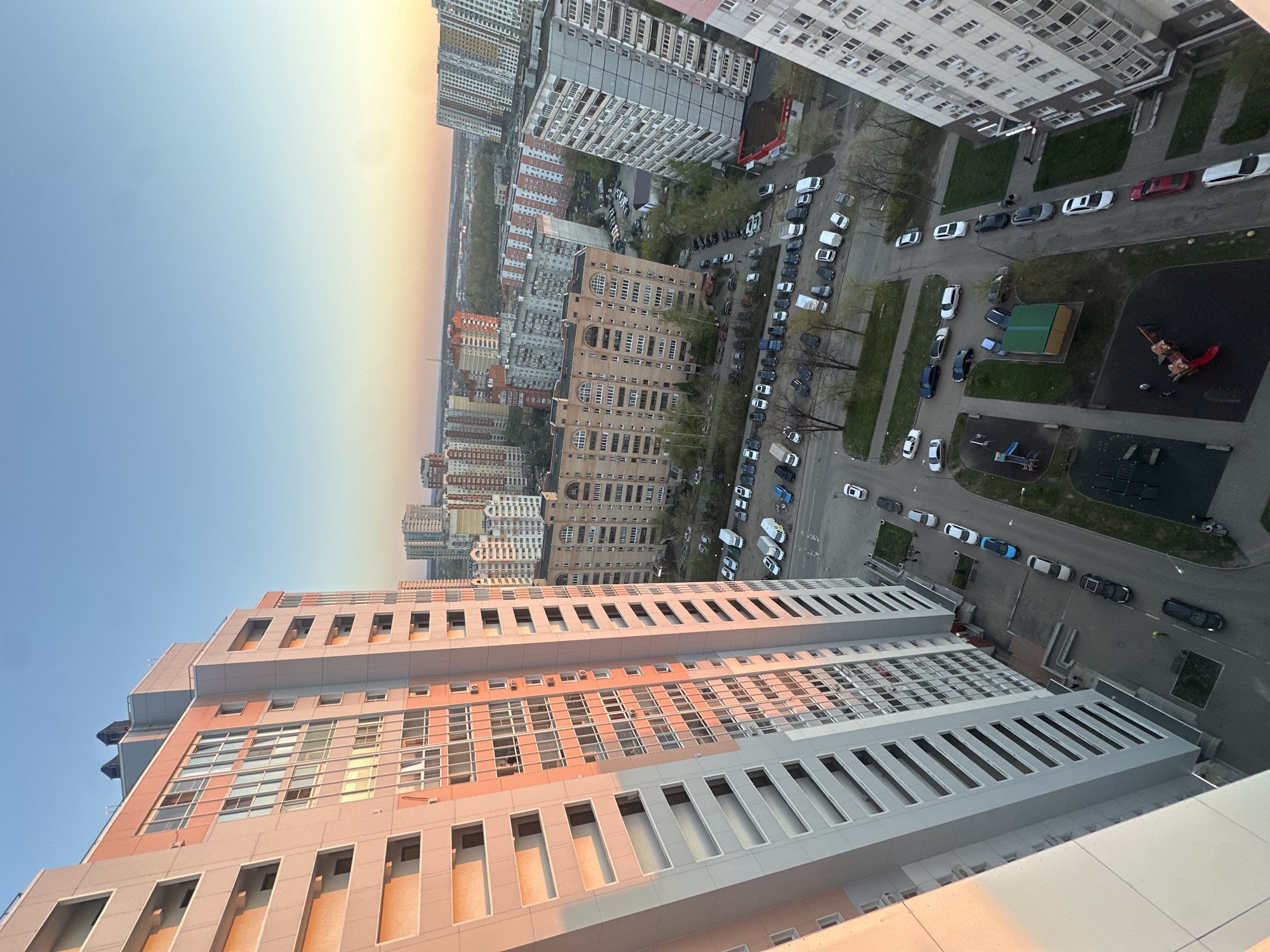 The image is a high-angle, daytime view of Khimki, Russia, capturing a dense urban landscape. In the foreground is a multi-story building with a distinctive architectural design featuring alternating rows of windows and recessed vertical panels. The building has a color palette of peach and gray.
Moving into the middle ground, the scene reveals a congested city environment. Numerous apartment buildings of various heights and architectural styles are present. Streets are visible, densely populated with parked cars of various makes and colors.
In the background, the image shows the further extent of the city fading into the horizon under a clear, blue sky. There appears to be a slight gradient or tint in the sky near the horizon, suggesting either a sunrise or sunset.
Overall, the scene depicts a typical urban landscape in Khimki, Russia, characterized by high-density residential buildings and a significant amount of vehicular traffic. There are no discernible people visible in the image. The environment appears to be relatively calm and orderly.
