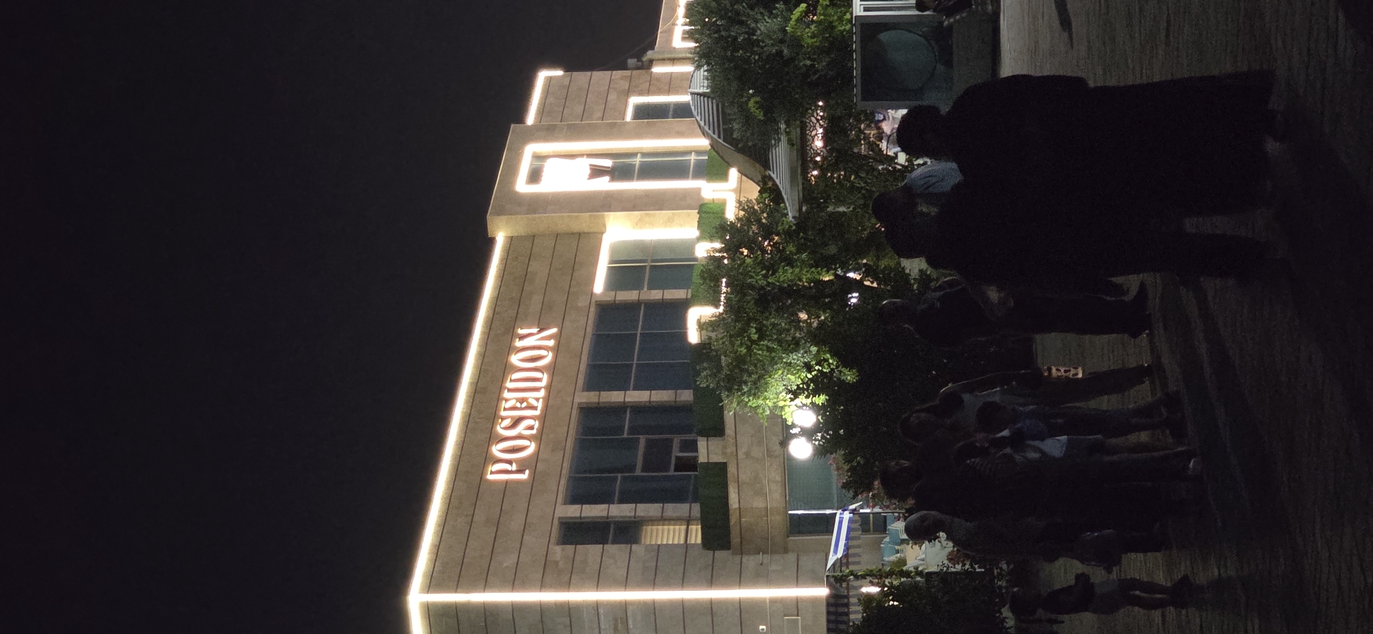 The image captures the exterior of a building at night, illuminated by exterior lighting and a prominent illuminated sign reading "POSEIDON". The building appears to be constructed of stone or a similar material and features large windows. In the foreground, a group of people are silhouetted against the lights, their features obscured by darkness. The scene suggests an outdoor gathering or event. The location is identified as Sumgayit, Azerbaijan.