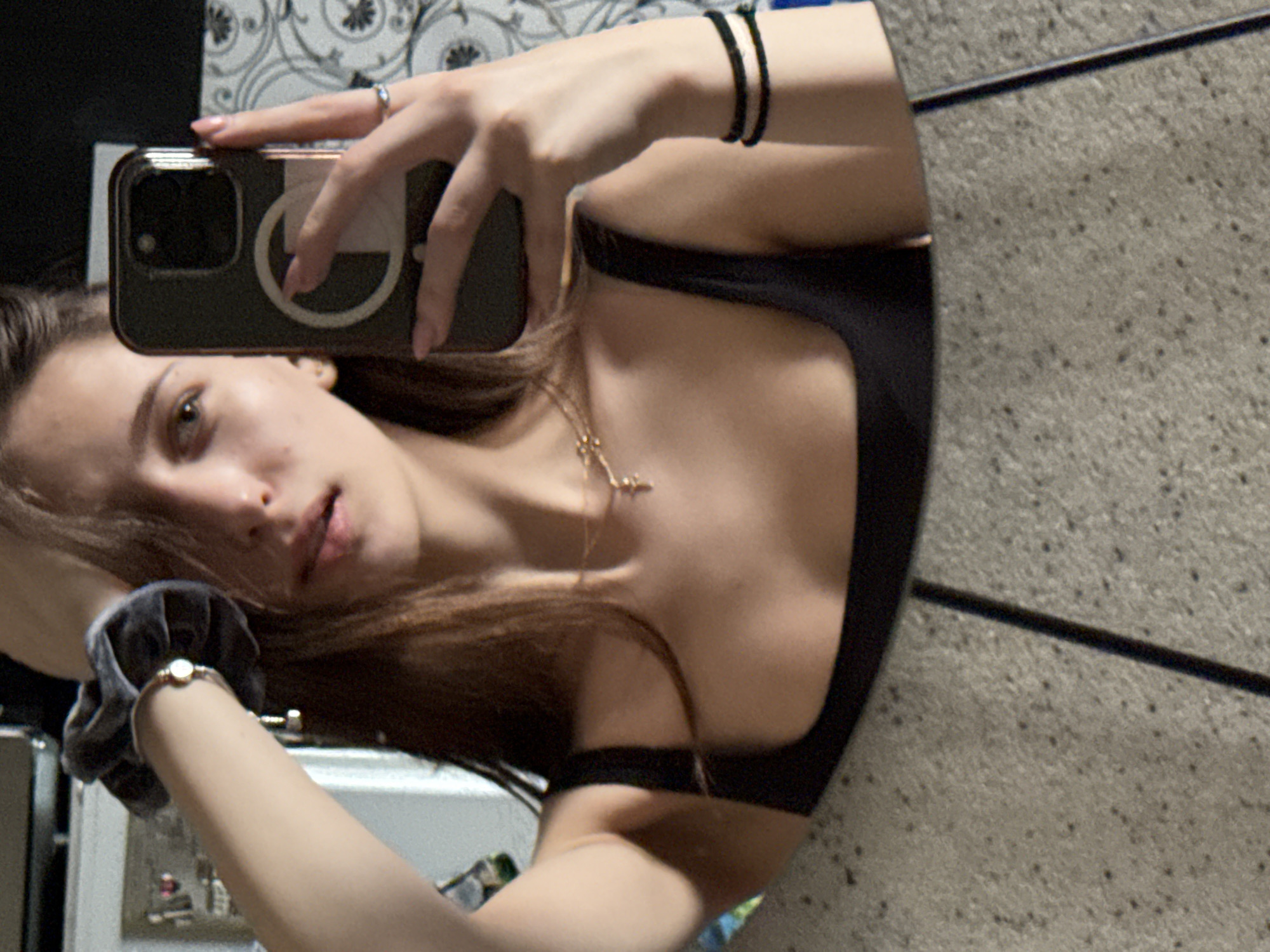A mirror selfie captures a young individual, likely female, with long brown hair, gazing towards the camera with a neutral and slightly pensive expression, her lips subtly parted. She is dressed in a black top or sports bra, complemented by a gold-colored necklace featuring a small cross-shaped pendant.
On her right wrist, she wears two thin black elastic hair ties, while her left wrist is adorned with a dark grey velvet scrunchie and a delicate bracelet holding a small, circular, light-colored charm. Her right hand, featuring a silver ring on the ring finger, holds a smartphone encased in a dark cover that displays a prominent white circular emblem, resembling a MagSafe ring.
The background, reflected in the mirror, reveals an indoor setting. To the upper left, a patterned wallpaper or fabric with a black and white floral or damask-like design is visible. To the right, a grainy, light grey wall surface with a distinct dark horizontal line is seen. The image is presented rotated 90 degrees clockwise, adding a dynamic angle to the composition. The time of day is not evident, but the setting suggests an interior space, consistent with a residence in Sofia, Bulgaria, as provided by the context. There is no discernible text within the image.