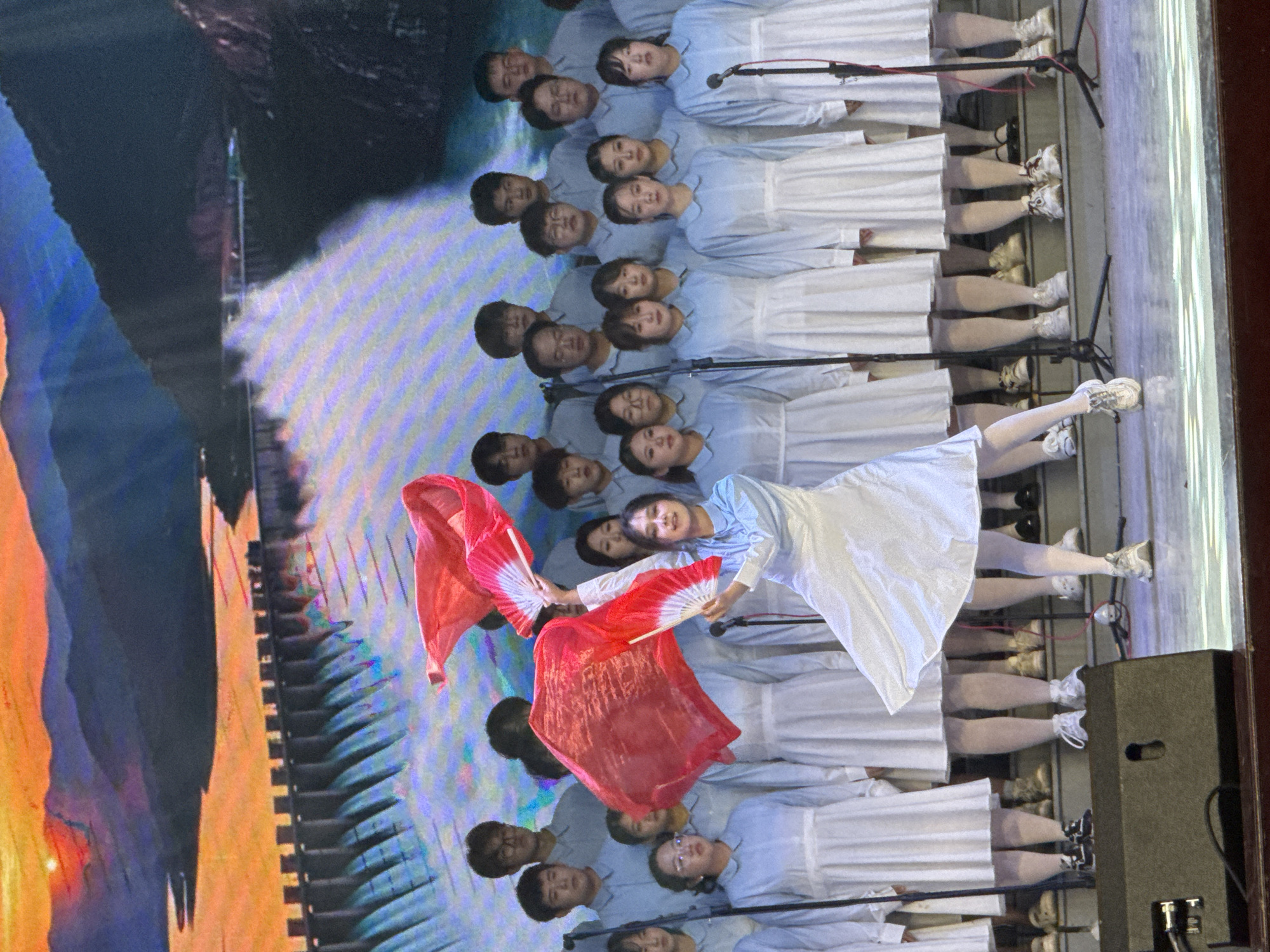 The image captures an indoor performance taking place on a stage in Guta, China. The primary subjects are a large group of young people, likely a choir or performance ensemble, arranged in multiple horizontal rows. They are uniformly dressed in light blue long-sleeved shirts paired with white skirts or dresses, often worn over white tights or leggings. Most performers are wearing white athletic sneakers, though some have black formal shoes. Microphones on stands are interspersed among the rows, indicating a vocal performance.
In the foreground and slightly to the right, a central female performer is actively engaged in a dance or movement. She is dressed similarly but distinctively holds two vibrant red fans or flags, one in each hand, which are unfurled and spread wide, adding a dynamic splash of color. Her expression is joyful, with a clear smile. The other performers maintain neutral or focused expressions.
The background is a large, brightly lit LED screen displaying an abstract and colorful projection featuring swirling patterns of blue, purple, orange, and yellow hues, which provides a dramatic visual backdrop. A dark speaker box is visible on the right side of the stage. The overall atmosphere conveys an organized and energetic cultural or artistic event. No discernible text is visible within the image.