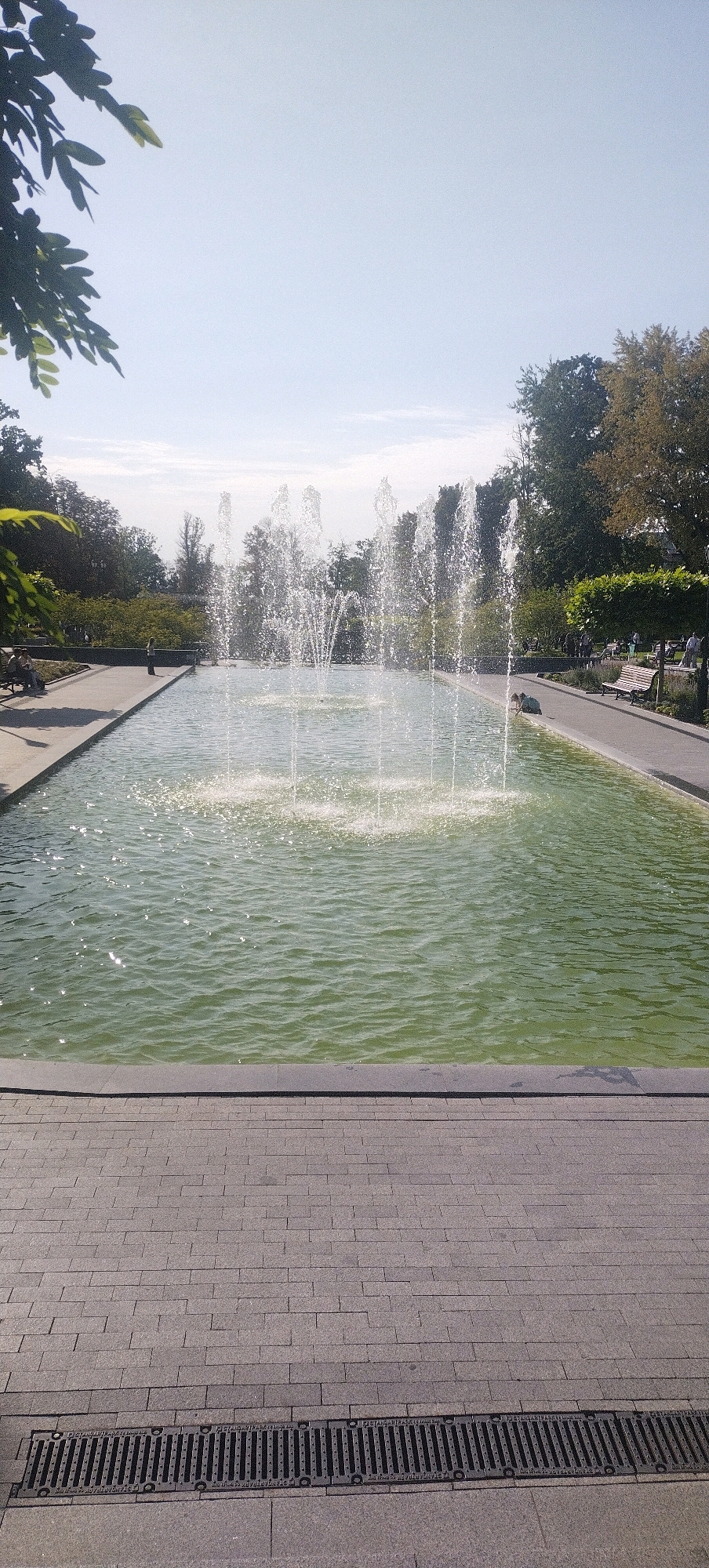 This vertical, eye-level shot depicts a bright, sunny day in Kharkiv, Ukraine, likely in a park or public square. The focal point is a rectangular fountain with multiple jets of water spraying upwards, creating a dynamic visual effect. The water is a greenish hue, and sunlight glints off its surface.
The fountain is bordered by a gray stone walkway and then a paved area made of gray rectangular bricks. A metal grate, likely a drainage channel, runs horizontally across the bottom of the frame, parallel to the brickwork.
To the left of the fountain, a concrete path runs alongside the water, with a few people seated on a bench, observing the scene. Further back, more trees and foliage create a lush green backdrop.
To the right of the fountain, a similar paved path is visible, with benches and more trees. Several individuals are scattered along this path, some standing and others seated. The sky above is a clear, pale blue with a few faint clouds. The overall atmosphere suggests a tranquil and pleasant outdoor environment.