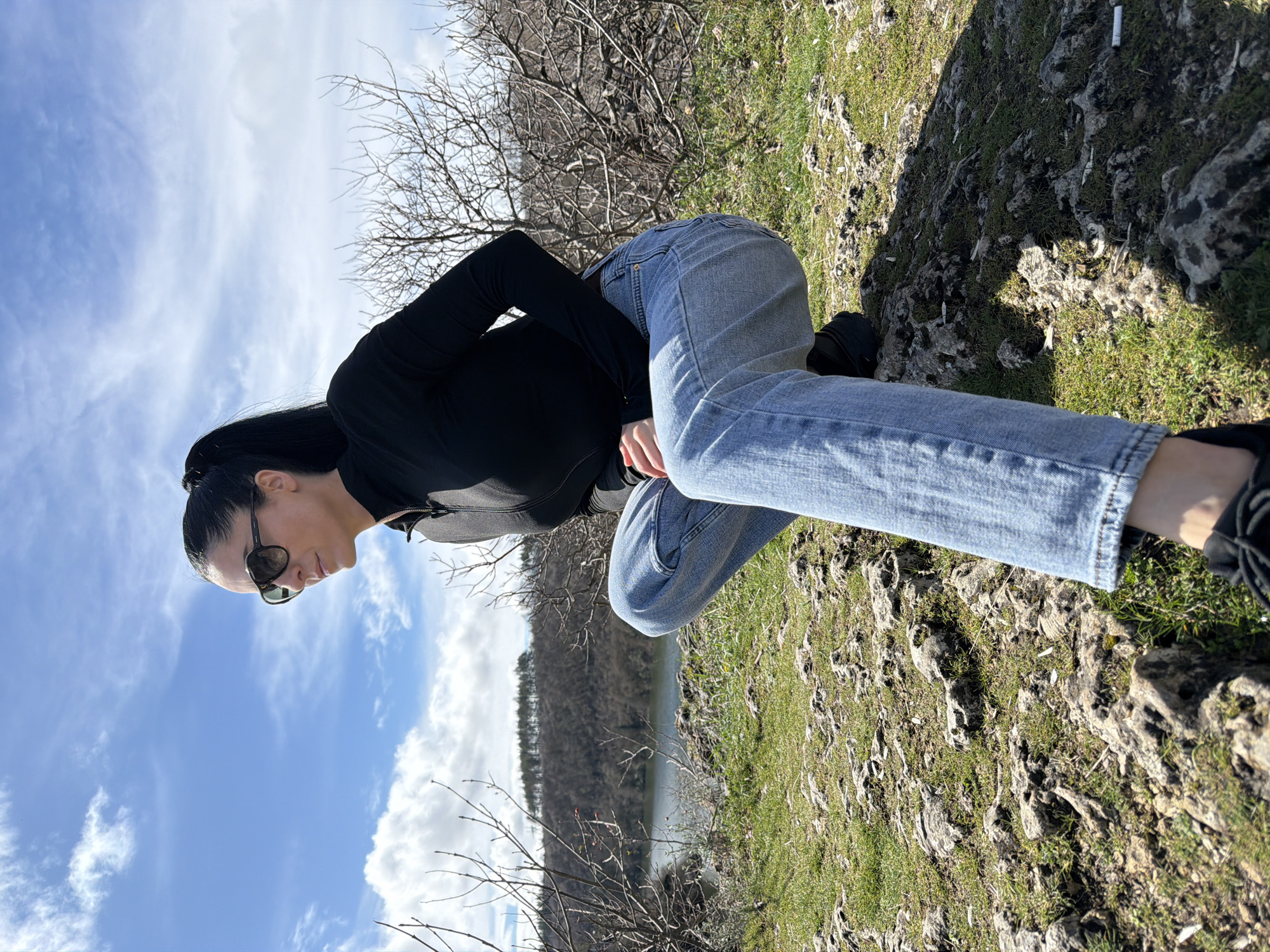 A female individual, with dark hair secured in a ponytail and wearing dark sunglasses, is observed in a crouched position on a rocky, grass-covered elevation. She is dressed in a black long-sleeved zippered top, light blue denim jeans, and dark athletic footwear. Her gaze is directed towards the left side of the frame, over the distant landscape, with her hands clasped in front of her. The immediate ground features exposed rocks with sparse green growth and several white cigarette butts scattered on the surface. Beyond the subject, the midground contains leafless deciduous shrubs and trees. A calm body of water, likely a river or lake, is visible in the valley, reflecting a bright, partly cloudy blue sky. On the opposing side of the water, a densely forested hill rises, topped with a distinct line of darker evergreen trees. The scene is consistent with the natural terrain found within Kaylaka Park, Pleven, Bulgaria.