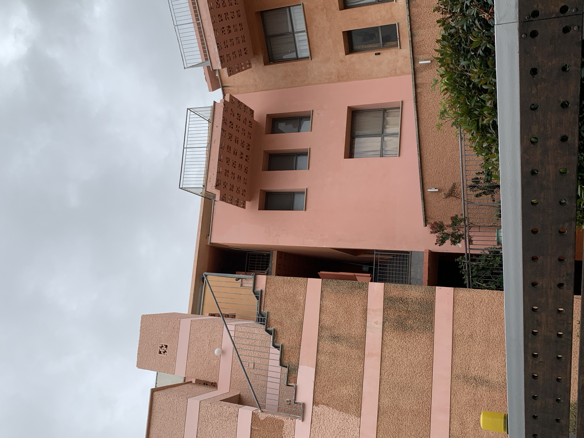 The image depicts an exterior view of a multi-story building in Tel Aviv, Israel. The architectural style incorporates textured peach-colored walls, with balconies and stairs visible. Windows are rectangular, and some have metal bars. Vegetation is seen growing near the building. The sky is overcast, suggesting a cloudy day. A metal structure with evenly spaced holes is visible in the foreground.