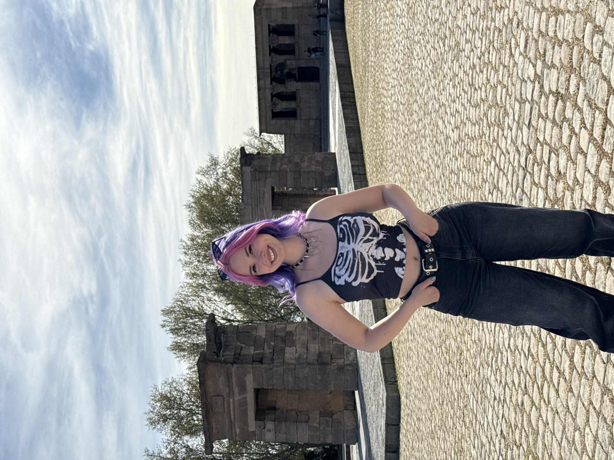 A woman stands in front of the Temple of Debod in Madrid, Spain. She is wearing a black tank top with a white skeleton design, dark jeans, and a studded belt. Her hair is dyed pink and purple. The ground is paved with cobblestones. In the background, the Temple of Debod is visible, along with some trees and a cloudy sky.