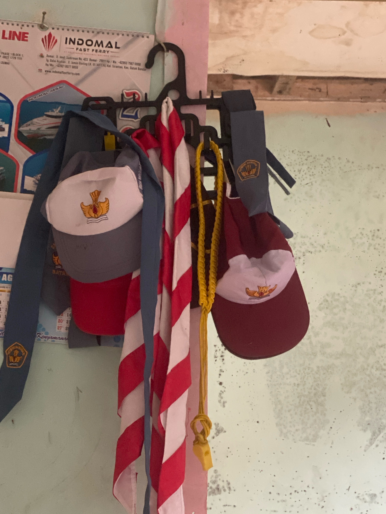 A close-up, eye-level shot captures a collection of scout uniforms and accessories hanging on a wall. The scene is dominated by the red and white striped scarf of a scout, a blue neckerchief, and two caps. One cap is gray with a white front panel featuring a yellow bird emblem, and the other is maroon with a white front panel also bearing the same emblem. A yellow whistle on a braided cord hangs from one of the caps. Behind the scout items, a poster or calendar is visible, featuring a ferry advertisement for "INDOMAL FAST FERRY" and listing contact details for locations in Dumai and Batam, Indonesia. The wall itself appears to be painted a light green color and shows signs of wear and mold, particularly on the right side of the frame. The lighting is somewhat dim, creating shadows and highlighting the textures of the objects and the wall. The overall impression is one of a storage space, possibly a classroom or common area, where these scout items are kept.