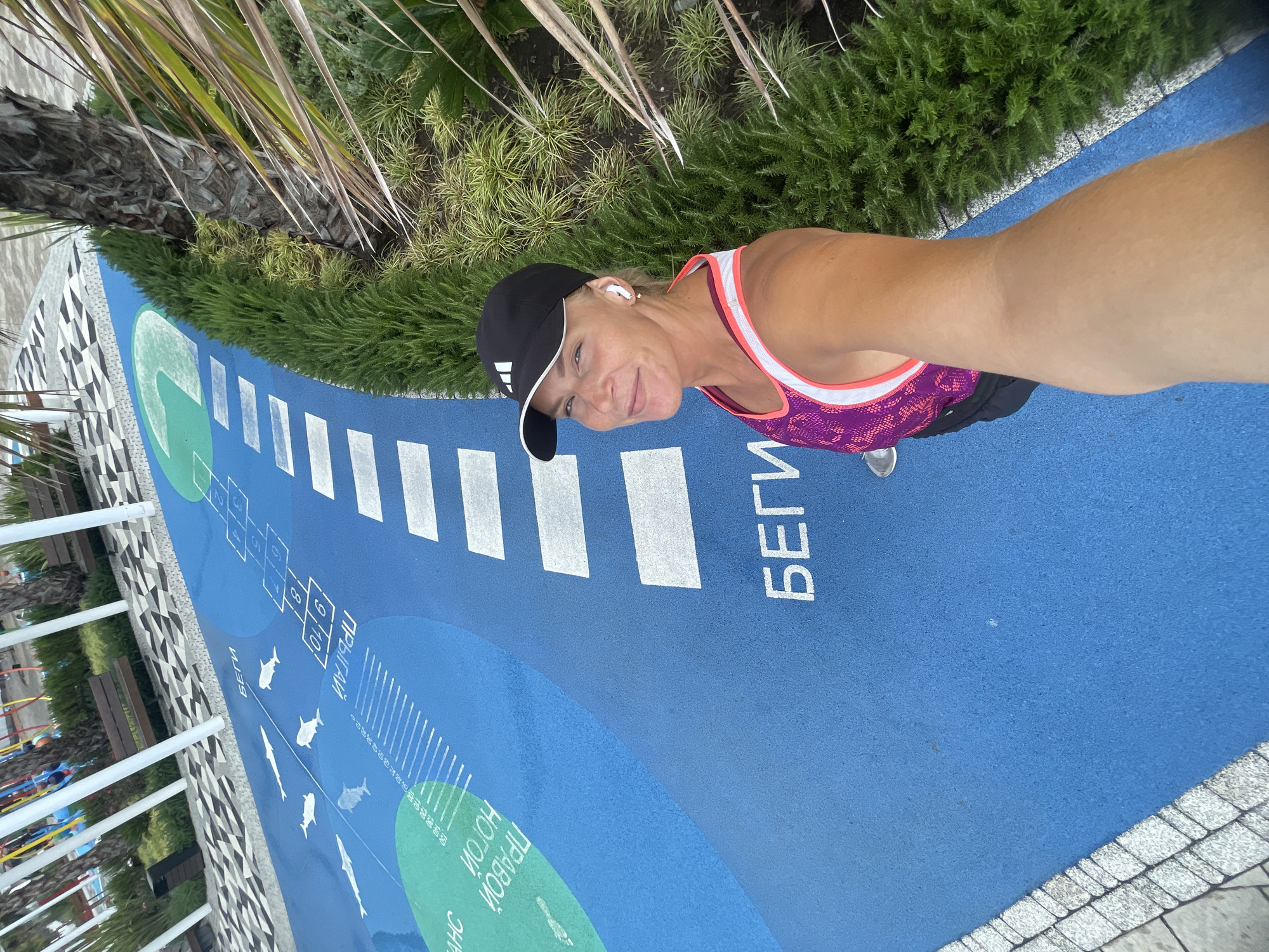 In a selfie taken at a Sochi, Russia park, a woman wearing a black Adidas cap and a purple and pink athletic tank top smiles at the camera. She has on white earbuds and is standing on a bright blue surface with white hopscotch squares and graphics of sharks and fish. To the right, lush green bushes and a palm tree are visible. The background shows patterned pavement, benches, and white poles, suggesting an outdoor recreational area. The text "БЕГИ" (Run) is visible on the blue surface.