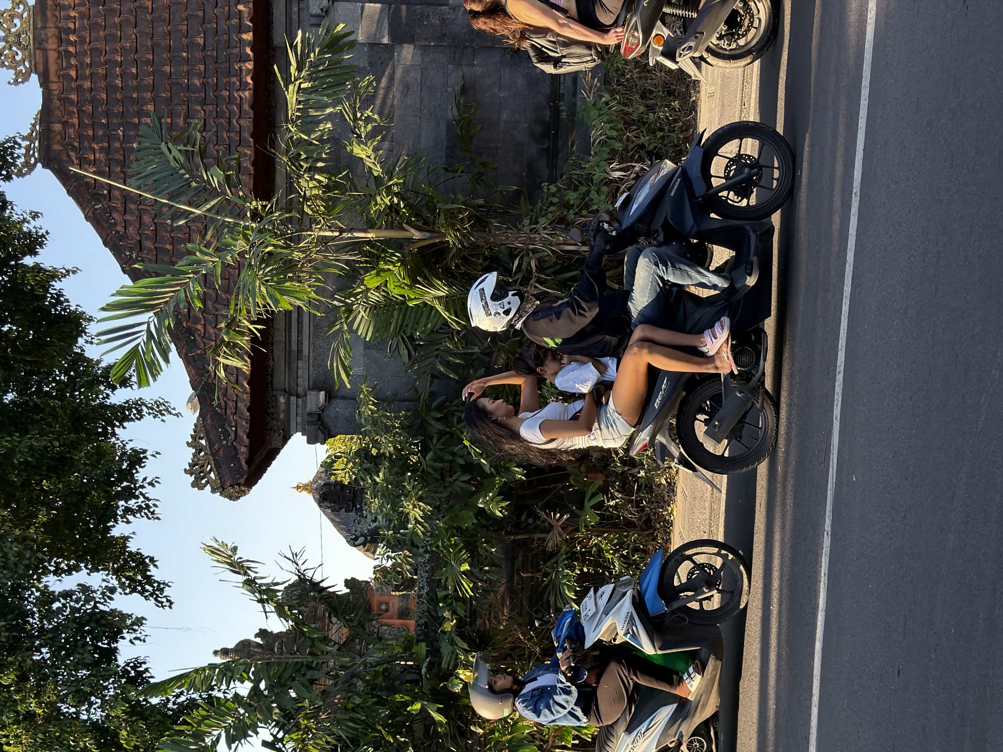 This detailed image captures a late afternoon street scene in North Kuta, Indonesia, featuring multiple motorbikes with riders and passengers. The setting is characterized by a clear blue sky, overhanging green tree canopies, lush tropical foliage including palm trees, and traditional Balinese architecture.
On the asphalt road, a solid white line is visible, and long shadows extend across the pavement from the left side, indicating low-angle sunlight typical of late afternoon.
From left to right:
1. A person wearing a grey full-face helmet, a light blue denim jacket over a white top, dark pants, and light-colored sandals is seated on a white and blue Honda Beat motorbike. The license plate "DV 492..." is partially visible. The rider is holding the handlebars and appears to be looking forward.
2. A dark-colored Honda Beat motorbike follows, carrying three individuals. The driver, a man, wears a white full-face helmet with dark markings, a black long-sleeved jacket, blue jeans, and dark footwear. A small child, wearing a white t-shirt, is positioned in front of the driver, between the handlebars and the driver's body. Behind the driver, a woman with long dark wavy hair wears a white short-sleeved top and light blue denim shorts, with light-colored sandals. Her right hand is raised, adjusting her hair.
3. On the far right, partially out of frame, the rear section of a third dark-colored motorbike is visible. A person, likely a woman, is seated on it, wearing a light-colored top (possibly pink or mauve) and carrying a black backpack.
The background behind the motorbikes consists of dense green tropical plants and trees, a dark grey stone wall constructed from rectangular blocks, and traditional Balinese structures. These include weathered buildings with ornate terracotta tiled roofs featuring decorative ridges and carved stone elements, along with dark, temple-like structures adorned with golden accents and intricate carvings.
