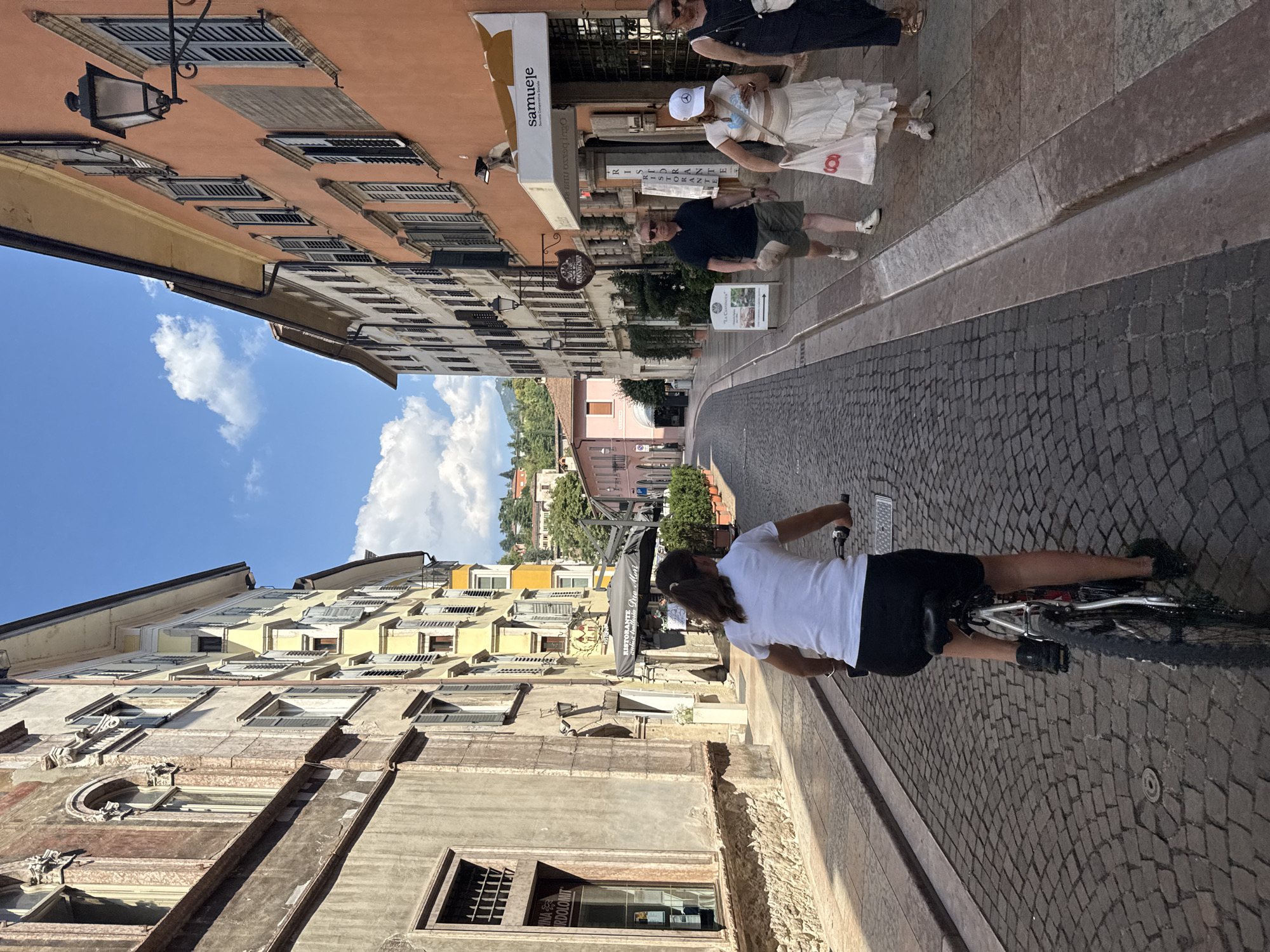 The image captures a street scene in Trento, Italy, likely a pedestrianized area given the cobblestone pavement and absence of cars. In the foreground, a young woman with dark hair pulled back in a ponytail rides a bicycle, wearing a white t-shirt and black shorts. She is viewed from behind and slightly to the side. Further down the street, three people are walking. A man in a dark shirt and shorts walks towards the viewer, holding a hat. Behind him, a woman in a white skirt and top, wearing a white cap, walks with a white shopping bag. To her right, another woman in a dark dress walks away from the viewer. The street is lined with buildings of varying architectural styles and colors. On the left, older buildings with stone bases and cream-colored upper floors feature arched windows and ornate details. On the right, buildings are painted in warmer tones, including yellow and orange, with numerous closed shutters. A sign with "samuele" is visible above a shop. The sky is blue with scattered white clouds. Greenery in potted plants adorns the sides of the street. In the distance, rolling hills covered with trees are visible.