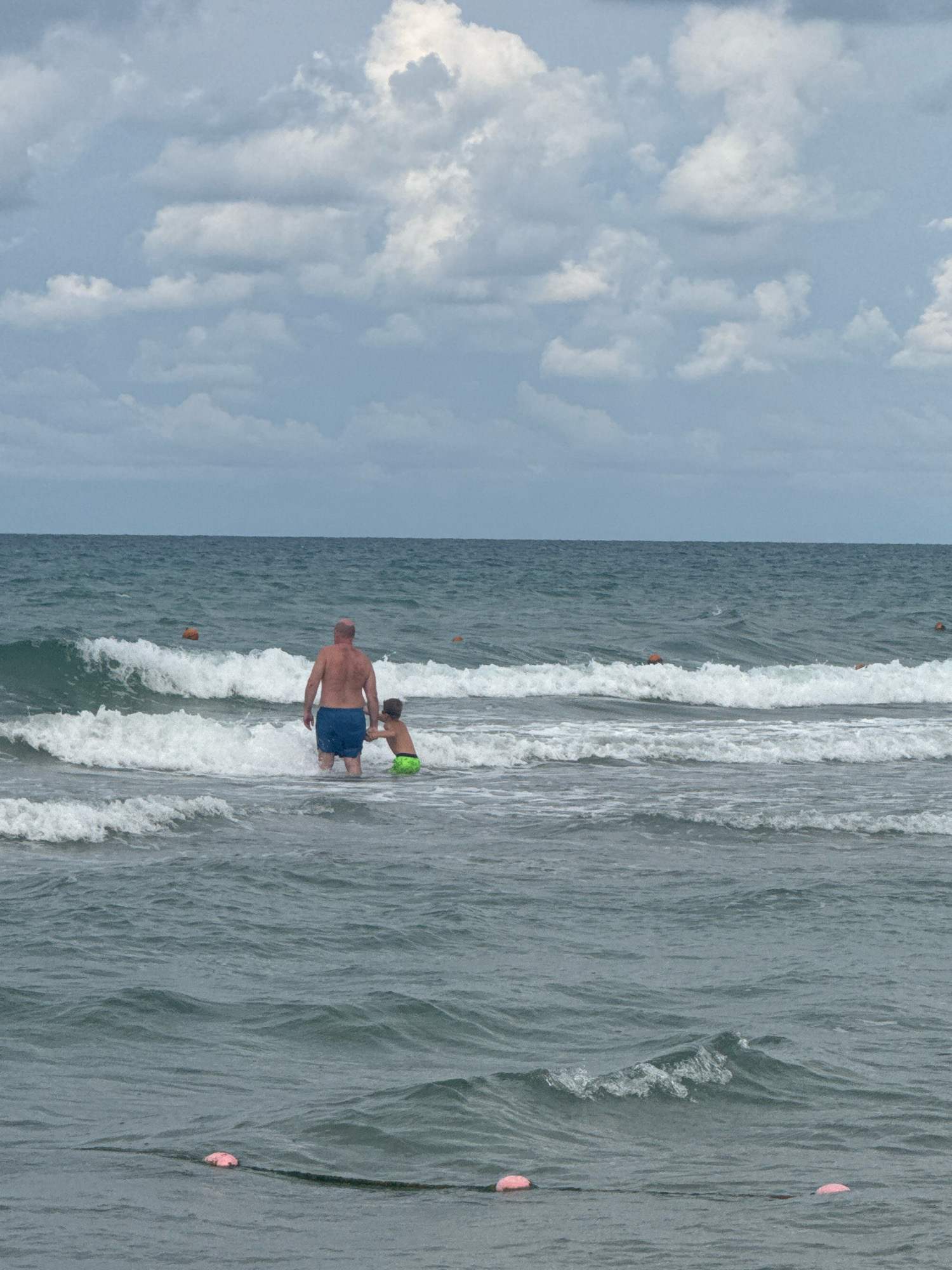 The image depicts a man and a child in the sea at a beach in Anapa, Russia. The man, who appears to be bald and is wearing blue swim trunks, stands facing away from the camera, with the water reaching his waist. Beside him, a young child in green swim trunks is also in the water, holding onto the man's hand. Gentle waves are breaking around them, and the sea extends to the horizon where it meets a cloudy sky. Orange buoys are visible in the water further out, suggesting a designated swimming area or marker. In the foreground, a rope with pink buoys floats on the water's surface. The overall scene conveys a calm, everyday moment at the beach.
