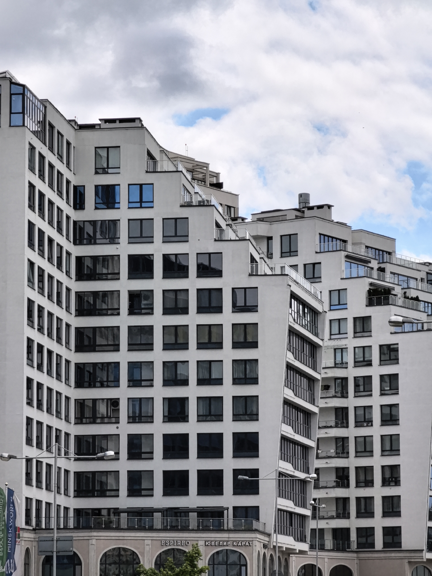 This is a medium shot, eye-level, vertical photograph of modern white buildings against a cloudy sky. The buildings have many windows and balconies, with a stepped architectural design. The sky is mostly gray with some patches of blue and white clouds. A street lamp is visible in the lower left portion of the image. Some flags are also visible in the lower left corner, with text that is not fully legible. The architecture suggests an urban setting.
