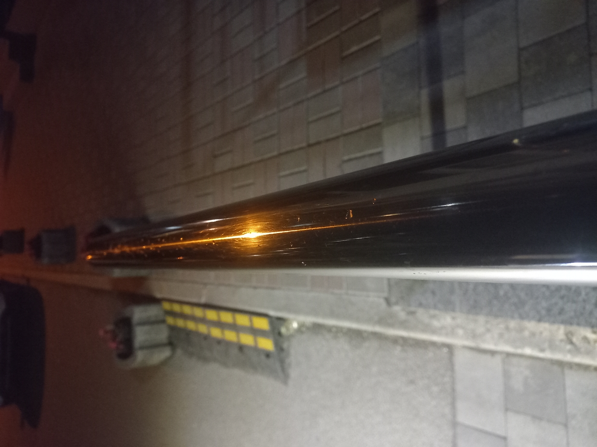 The image shows a close-up view of a reflective metal railing. Below the railing, there's a section of textured sidewalk pavement, partially visible. A yellow and black rubber parking bumper is also visible. In the background, blurred, a wall with rectangular blocks can be seen. The setting appears to be an urban streetscape at night. The photo does not provide enough information to identify the exact location within Kyiv.