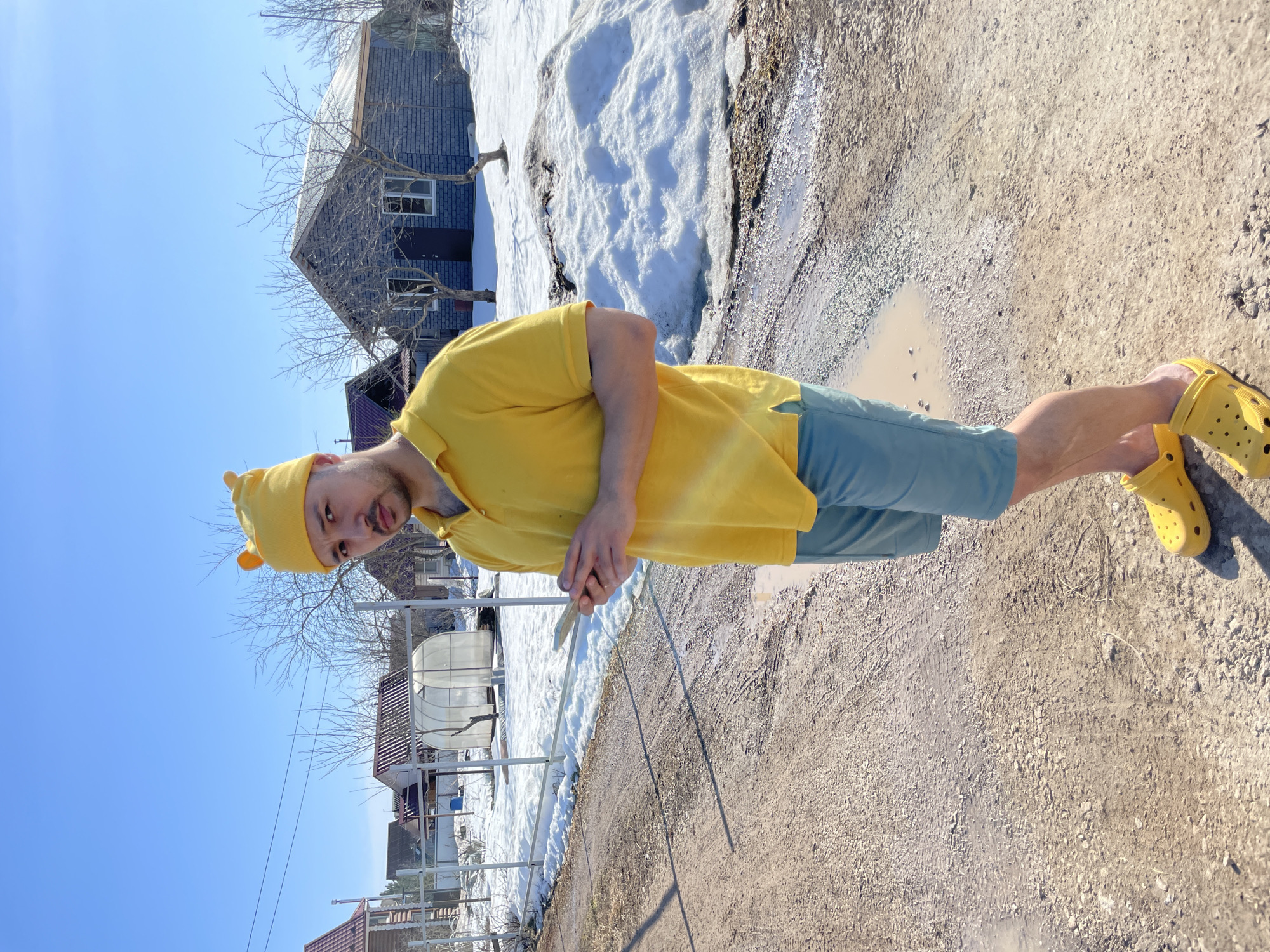 A man stands on a dirt road in Zav'yalovo, Russia. He wears a yellow shirt, blue shorts, yellow Crocs, and a yellow hat with bear ears. In his hands, he holds a thin metal rod. Snow patches remain in the background near a gray brick house. A white greenhouse is visible behind a metal fence. The sky is blue and clear. A puddle lies on the road near the man's feet.