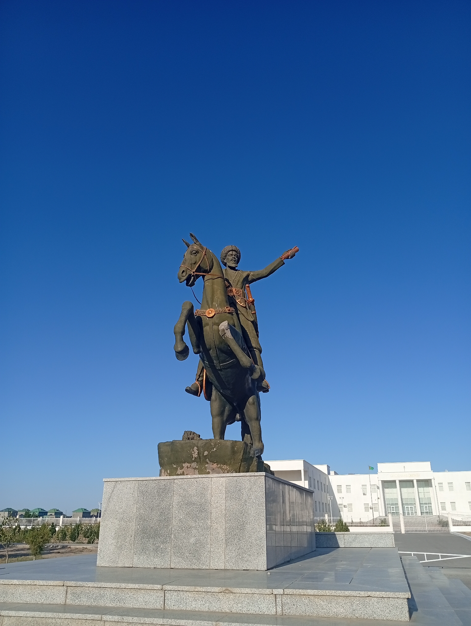An equestrian statue stands prominently against a clear blue sky. The bronze statue depicts a man in traditional attire riding a horse rearing on its hind legs. The man, with a beard and a fur hat, has his right arm extended forward, as if gesturing or pointing. He wears a tunic with ornate gold detailing and a sword at his side. The horse is adorned with decorative leather straps. The statue is mounted on a multi-layered pedestal, the lower portion of which is made of granite and the upper portion of weathered concrete, partially covered in moss or debris.
In the background, to the left of the statue, are low-rise buildings with green roofs, surrounded by trees and a fence. To the right of the statue, a modern, white multi-story building with a symmetrical facade and numerous windows dominates the background. A flag, possibly of Turkmenistan, flies from a pole on this building. The foreground consists of a paved area and stone steps leading up to the pedestal. The overall scene is set outdoors, under bright daylight, suggesting an open public space. The statue is likely a monument dedicated to a historical figure. Based on the architectural style and the presence of a flag that resembles the national flag of Turkmenistan, the location is identified as Seýdi, Turkmenistan.