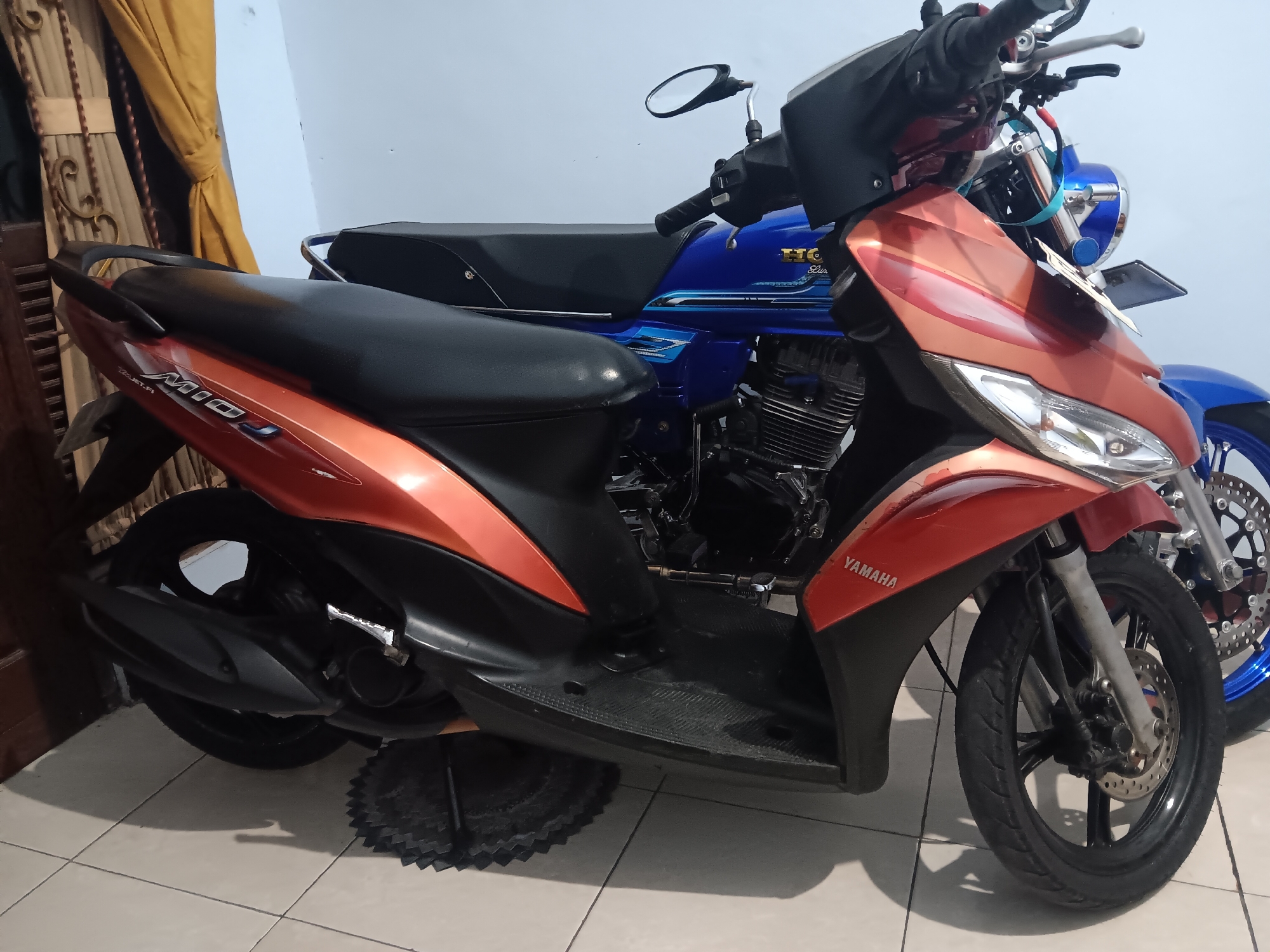 In Batursari, Indonesia, two motorcycles are parked side-by-side on a tiled floor. The motorcycle in the foreground is a Yamaha Mio, with an orange and black color scheme. It is a scooter-style motorcycle. Behind it is a blue Honda motorcycle, possibly a classic or retro style with a visible engine and chrome accents. The motorcycles are parked indoors, with a plain light-colored wall and a curtain visible in the background. The lighting suggests an indoor setting, possibly a garage or a covered parking area. There are no people visible in the image. The scene is static and focuses on the parked vehicles.