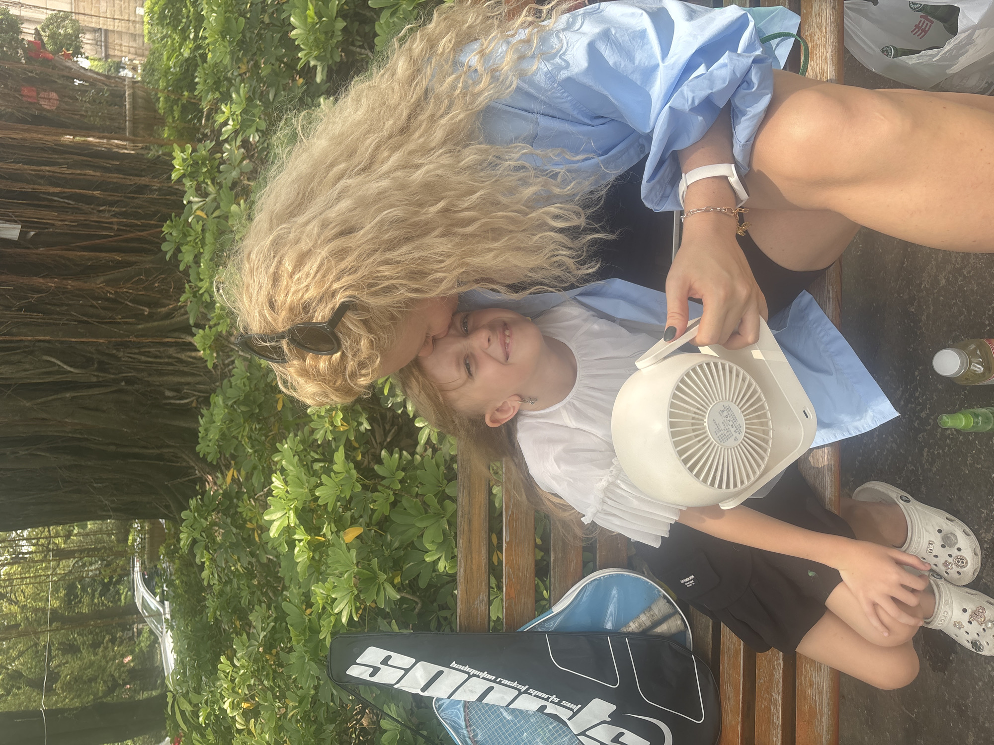 In Nanhai District, China, a woman with long, curly blonde hair kisses a young girl on the forehead while both are seated on a wooden bench. The girl, who has blonde hair and is wearing a white ruffled shirt and black shorts, holds a white portable fan. A tennis racket bag is visible to the left of the girl. The background features lush green foliage and the large, gnarled roots of a tree.
