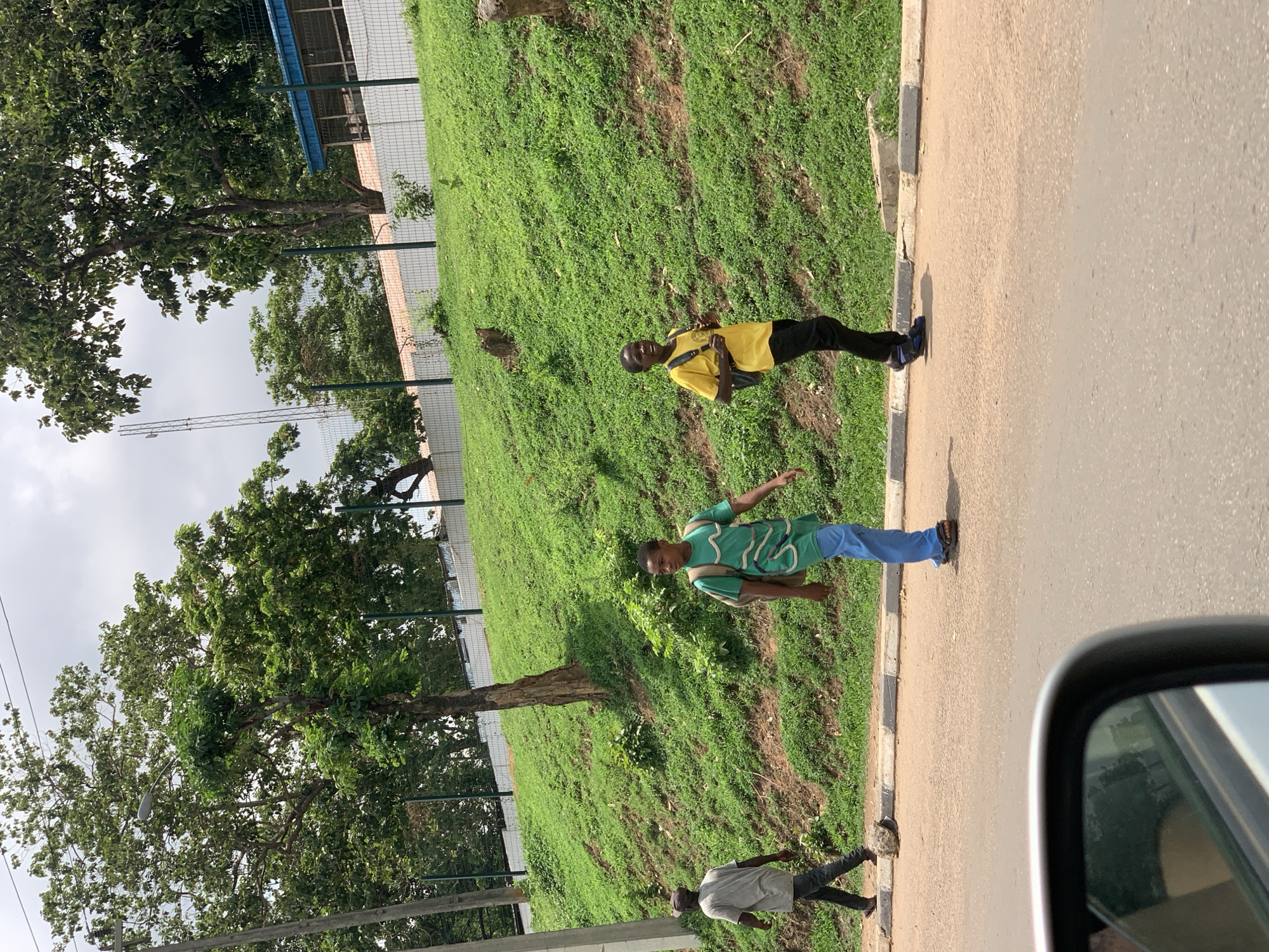 The image shows a street scene in Nyanya, Nigeria. There are three people visible: one wearing a yellow shirt, one wearing a green shirt, and one wearing a grey shirt. All three are located on the sidewalk bordering a grassy, inclined embankment. The yellow-shirted individual is standing still. The green-shirted individual is standing with their arm extended. The grey-shirted individual is walking. A road is next to the sidewalk, and the image seems to have been taken from a vehicle. There is a fence separating the embankment from what seems to be a building or a facility. Trees are present in the background. The sky is overcast.