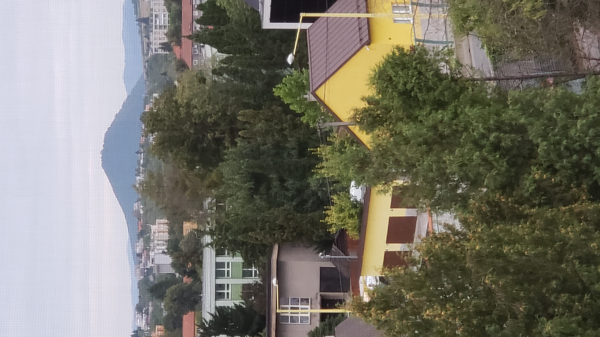 The image shows a view from an elevated position, likely a window, looking out over a suburban or semi-urban landscape. In the background, a distinct, rounded mountain dominates the skyline under a cloudy sky. Closer in, a range of buildings, including apartment blocks and smaller houses, are nestled amongst dense green trees. In the foreground, a yellow-painted building with a brown roof is visible, featuring garage doors. Adjacent to this is a darker, grey building with windows and a prominent yellow lamppost. The scene is characterized by a mix of residential architecture and abundant vegetation, with the prominent mountain serving as a backdrop. The image appears to be taken in the city of Prešov, Slovakia, with the mountain likely being the Zemplínské vrchy or a similar prominent geographical feature in the region. There are no people visible in the image.
