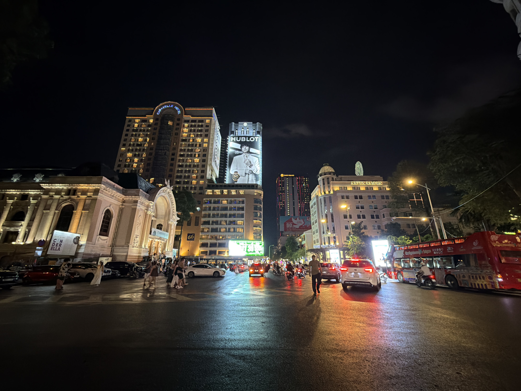 The image shows a street scene at night in Thủ Đức, Vietnam. The street is wet, reflecting the lights of the city. There are several buildings visible, including the Caravelle Hotel, a building with a large Hublot advertisement, and the Saigon Opera House. There are many pedestrians on the sidewalks and crossing the street, as well as cars, motorcycles, and a red double-decker tour bus. Streetlights illuminate the scene.