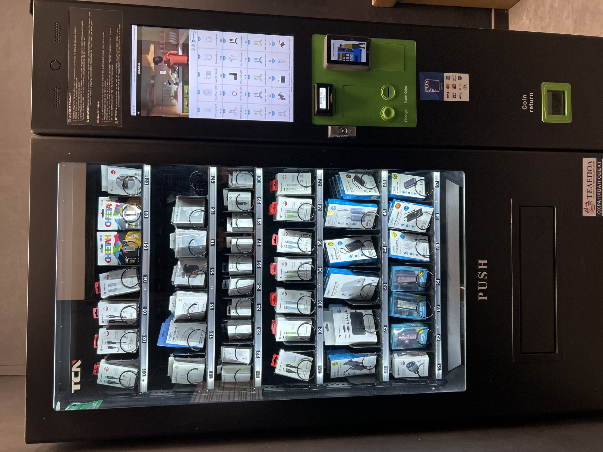 Here's a detailed description of the image:
The image depicts a vending machine stocked with electronics and accessories. The vending machine is black and silver, with a clear front panel displaying the products available. The panel is organized into columns and rows, each containing a different item such as charging cables, power banks, and memory cards. Each slot is labeled with a product code (e.g., '01', '02', '03') and price (e.g., '€15', '€22', '€20').
Above the product display is a control panel. It features a touchscreen interface displaying product options and payment instructions in Bulgarian, and English, along with a small payment screen. Additional features include slots for inserting coins, a change dispenser, a card reader, and a coin return. The machine accepts contactless payments via VISA, Pay, and G Pay.
A sticker on the lower right side of the machine displays the logo of "ТЕЛЕПОЛ", indicating a protected object, further suggesting a location in Bulgaria. A small sticker on the machine’s front panel reads “TCN”.
The instructions are displayed in both Bulgarian and English. The instructions detail the payment process, including selecting a product on the touchscreen, contactless payment at the terminal, and retrieving the product from the tray below.
Based on the "ТЕЛЕПОЛ" sticker, the photo was likely taken in Bulgaria. Without additional context, the specific location within Veliko Tarnovo cannot be determined, but it is situated within the context of a supervised area.