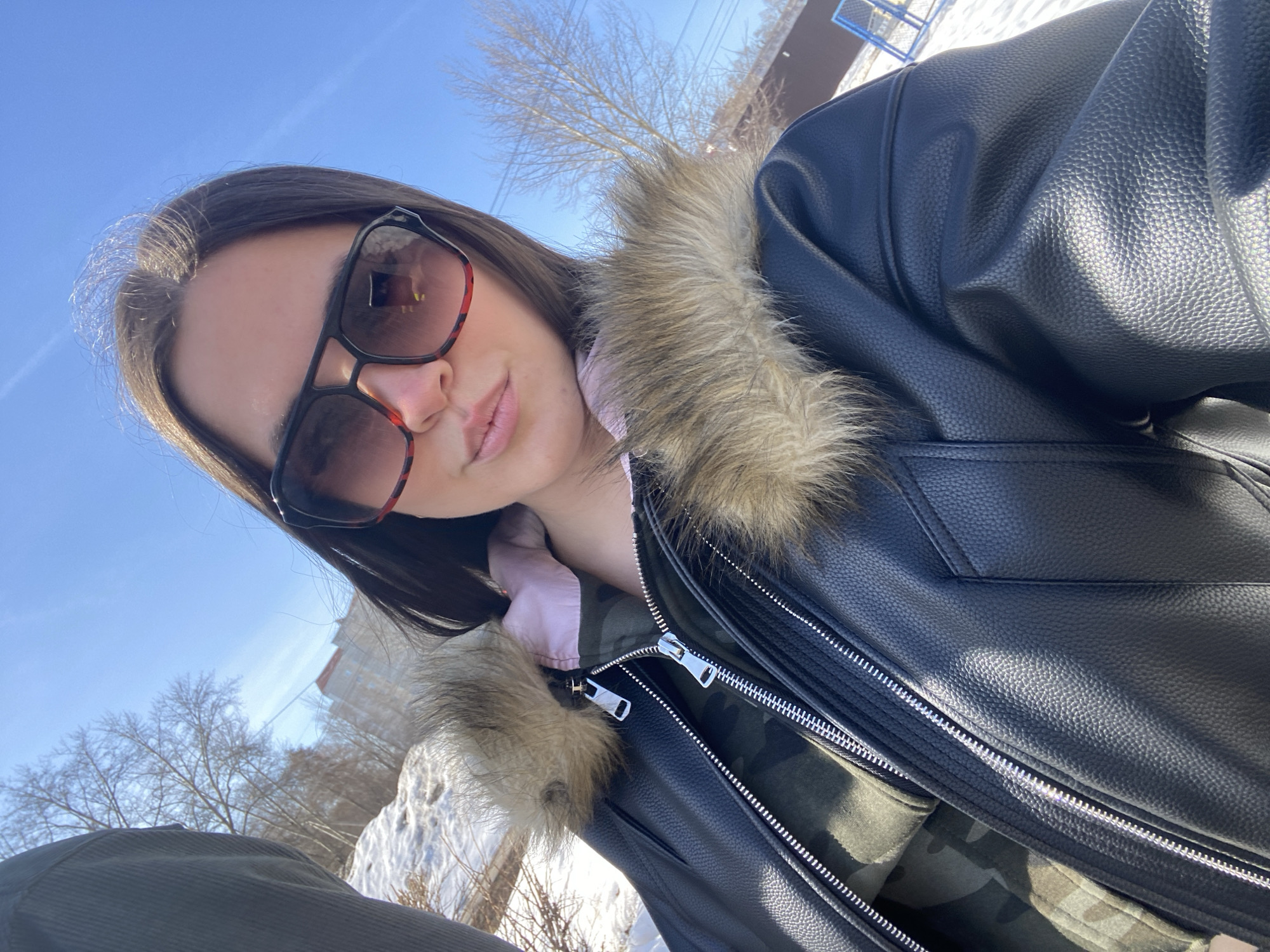 The image shows a young woman taking a selfie outdoors. She is wearing sunglasses and a dark leather jacket with a fur-lined collar. Underneath, she appears to have a pink shirt and a camouflage-patterned jacket. The background reveals a bright blue sky, bare trees, patches of snow, and a building in the distance. Based on the location tag, this image was taken in Chaikovsky, Russia.
